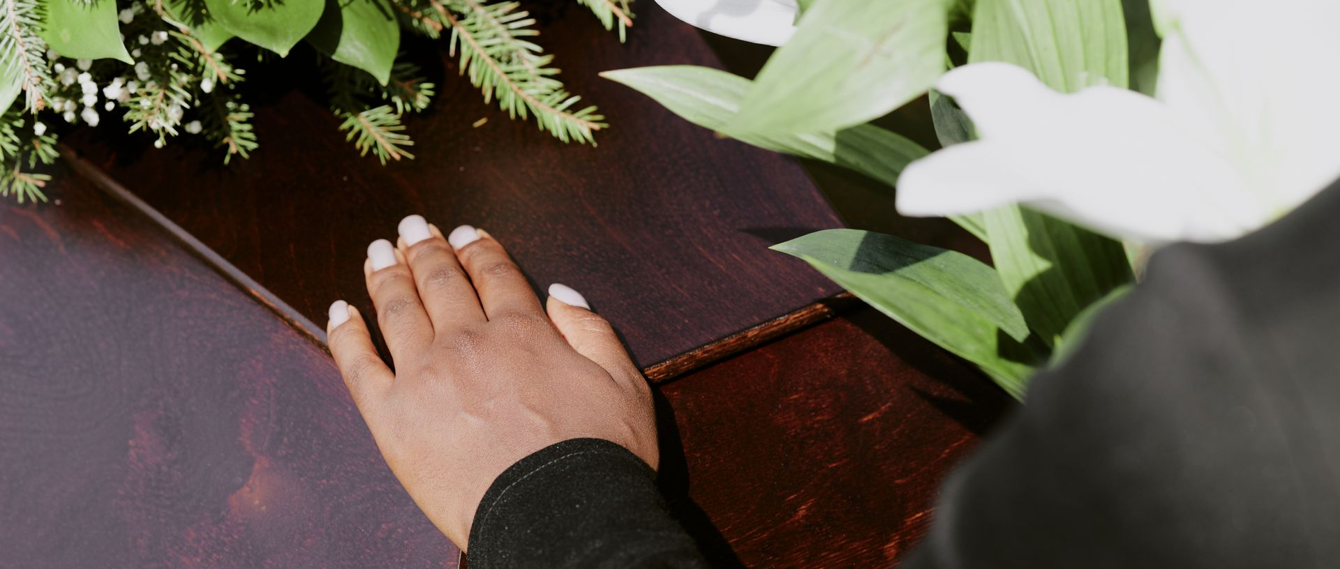 Hand resting on a dark wooden coffin beside white flowers and green foliage