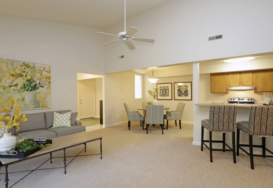 Living room with couch, dining table, bar stools, and a painting. Beige walls and carpet.