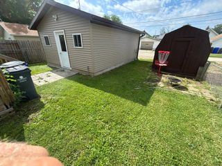 Backyard with green lawn, beige shed, dark storage shed, and garbage bin under a partly cloudy sky