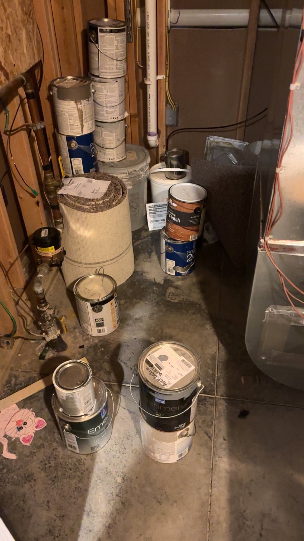 Paint cans and buckets stacked in a cluttered utility area with a concrete floor and wall pipes.