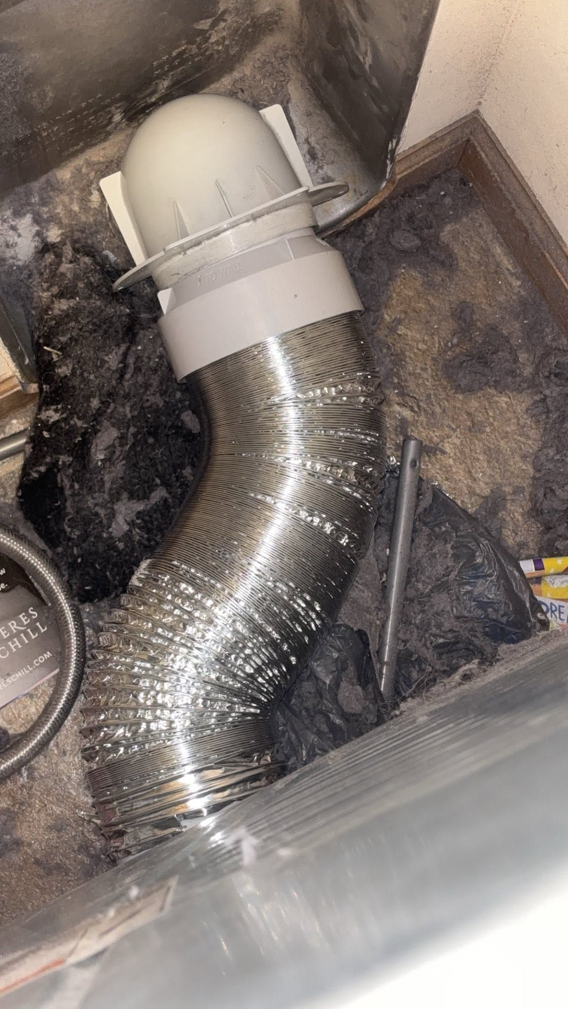 Flexible silver duct hose attached to a white vent in a dusty crawlspace beneath a home