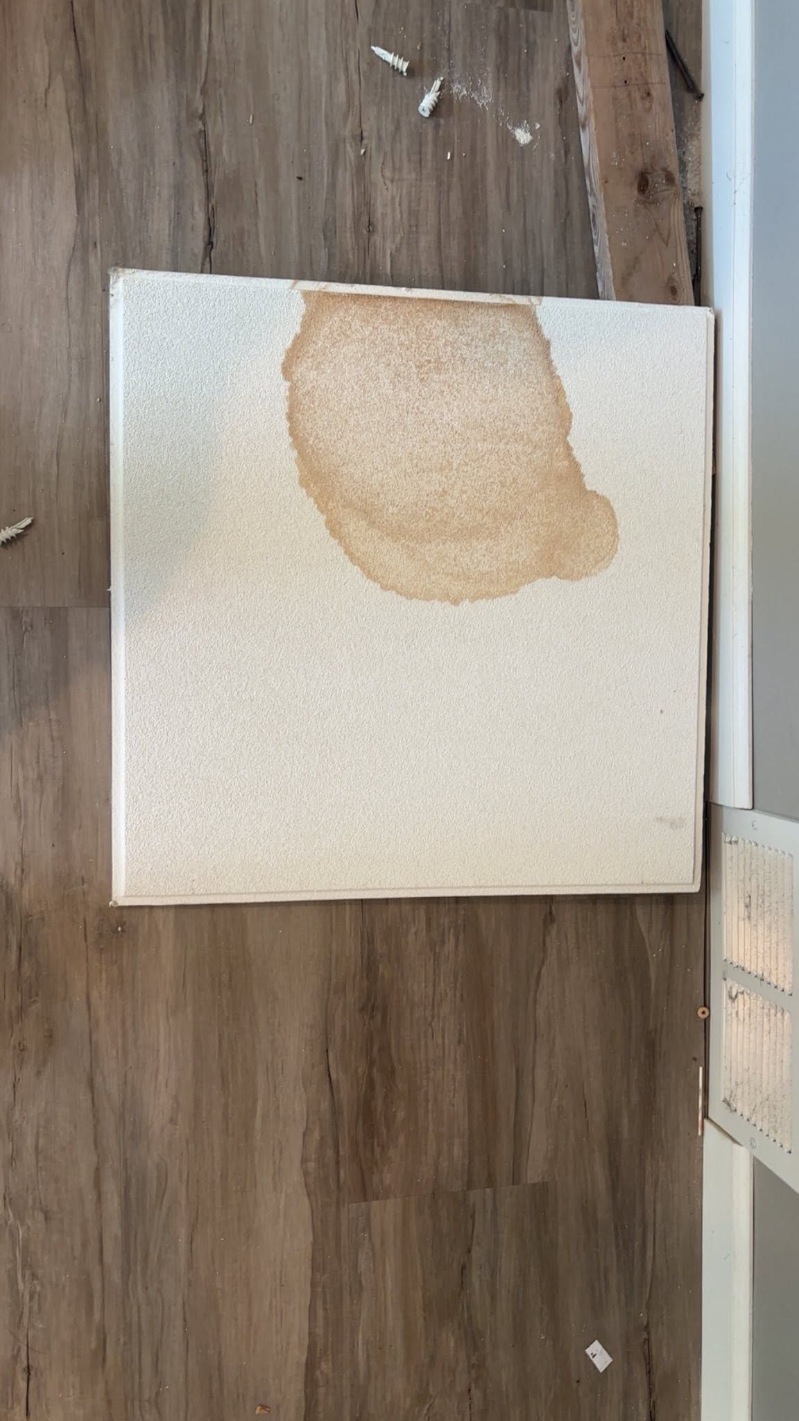 White square tile with a large brown water stain on top, lying on a wood floor.