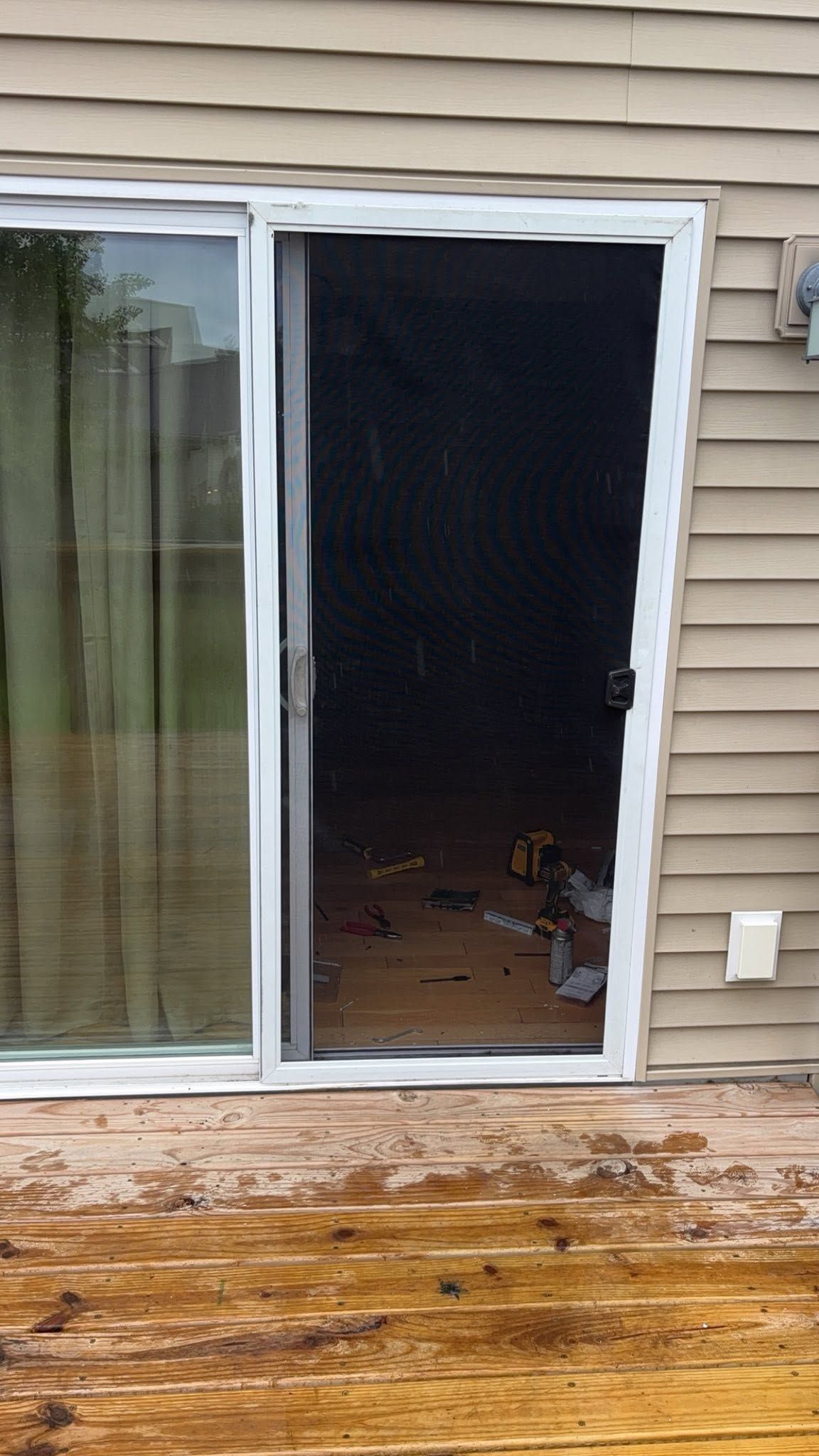 Open sliding glass door to a dark room, with wet wooden deck outside and beige siding.