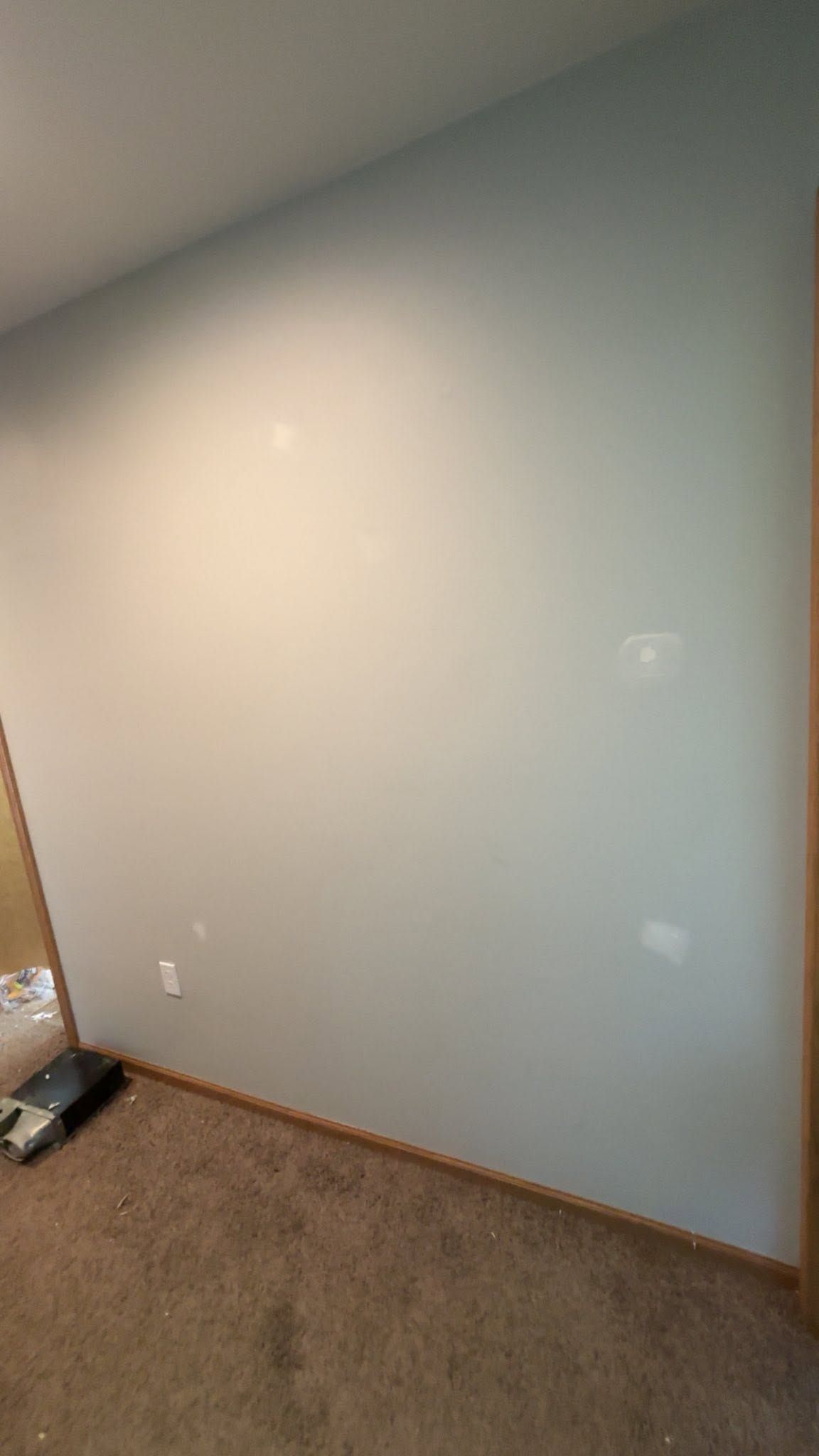 Empty beige wall above carpeted floor in a room, with white outlets and trim visible on the left.