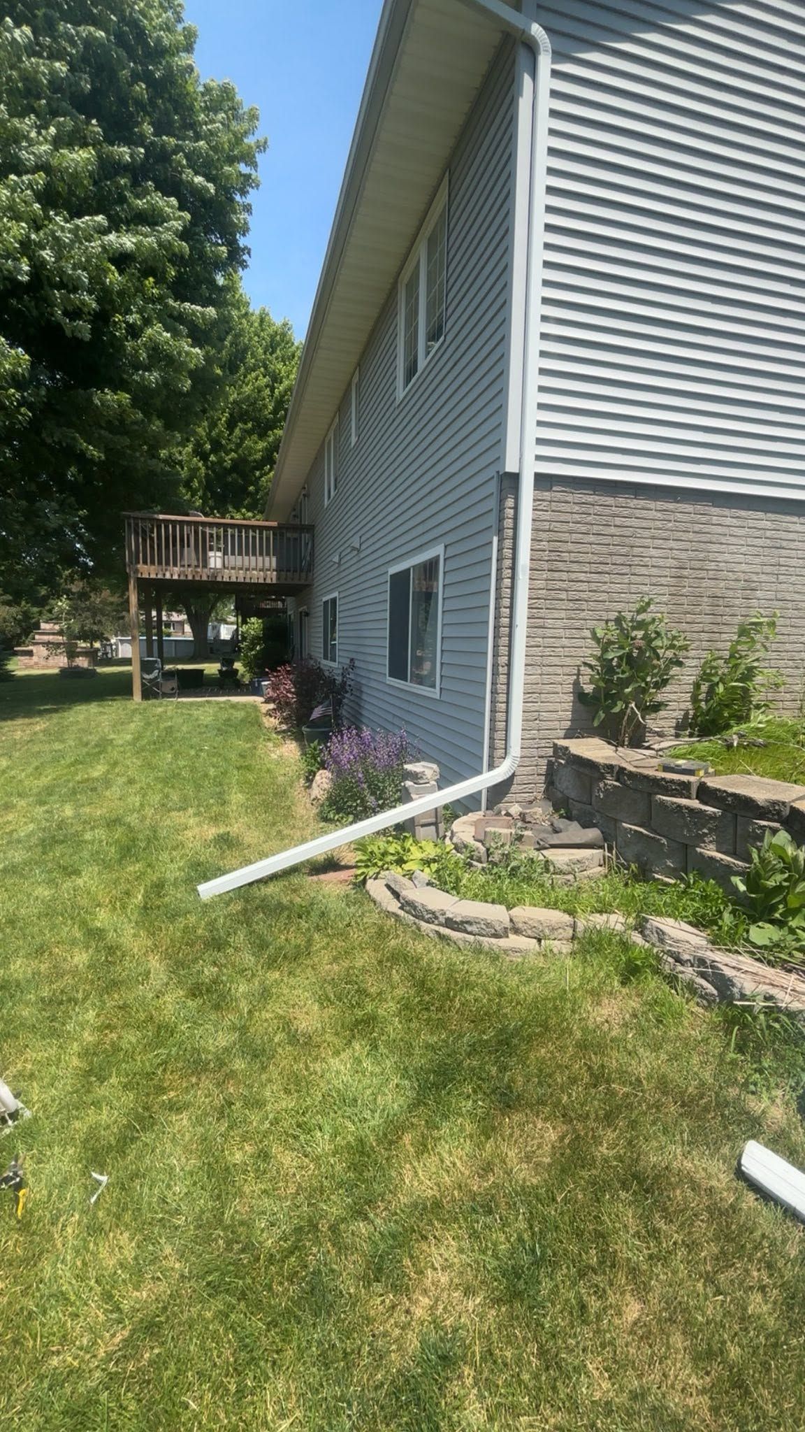 Side of a gray house with stone foundation, green lawn, white downspout, and a small deck in the background