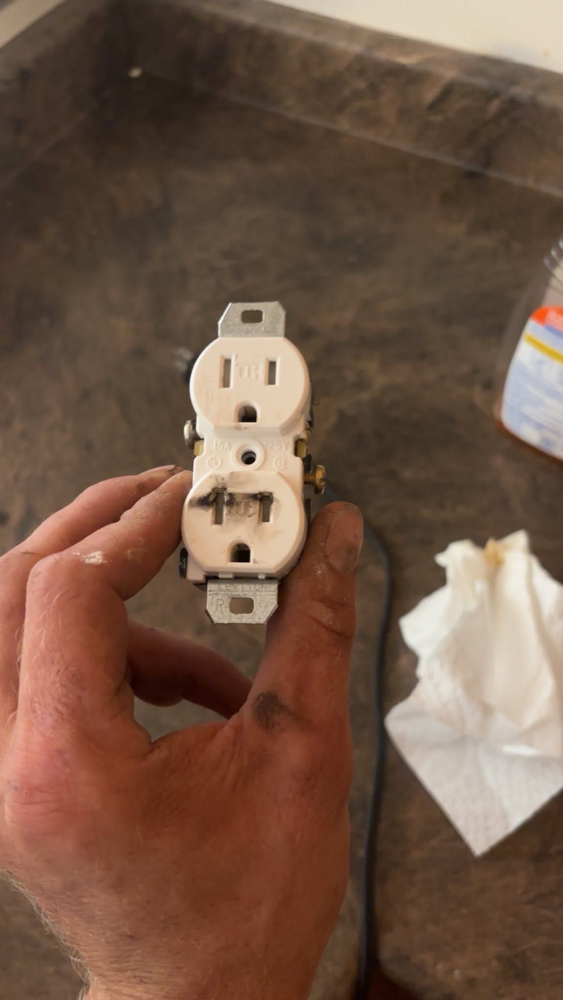 Hand holding a white electrical wall outlet over a brown countertop, with a tissue nearby.