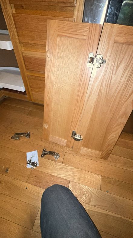Broken wooden cabinet door on hardwood floor with detached hinges and screws scattered nearby.