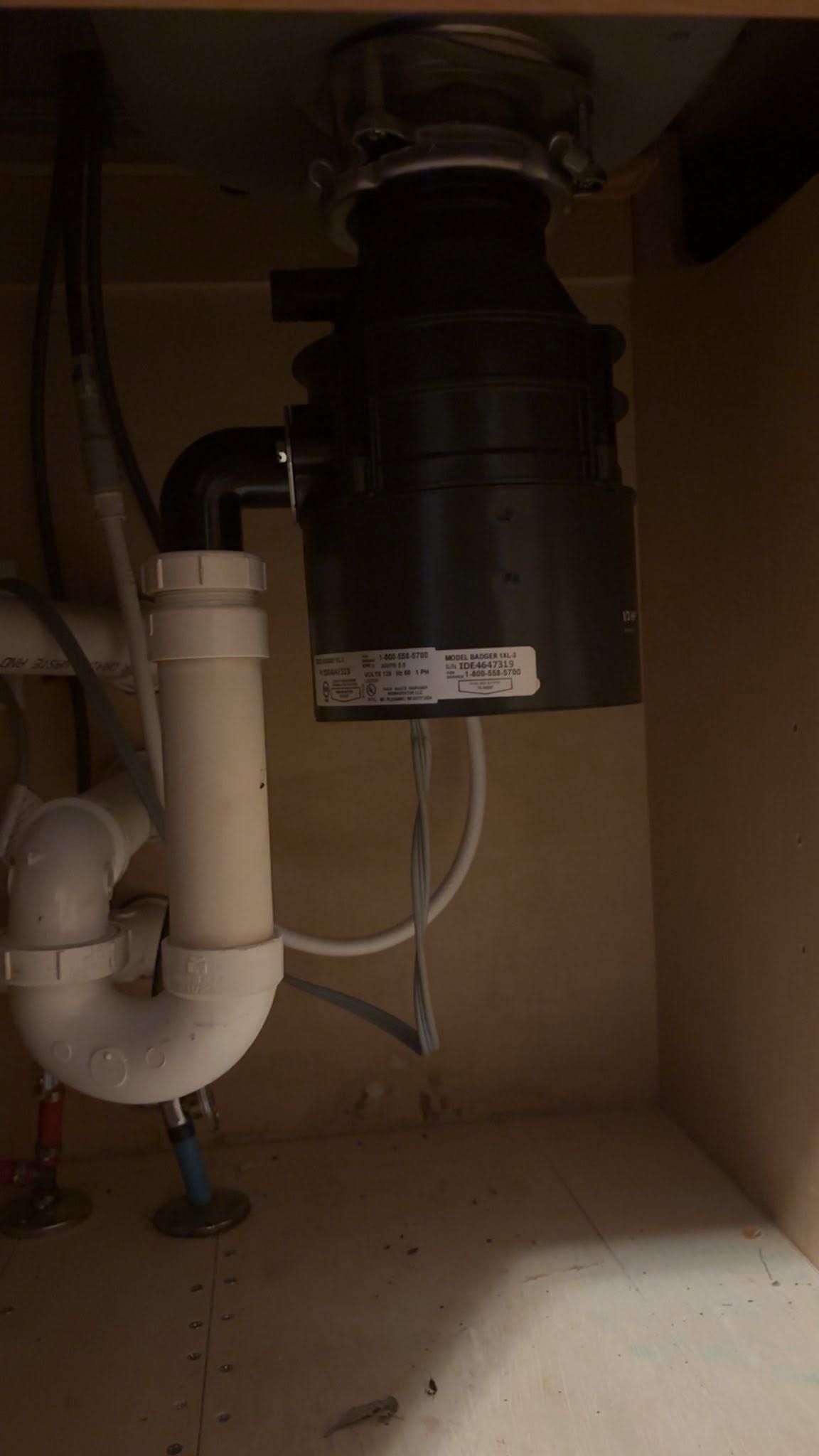 Under-sink plumbing with white pipes and a black garbage disposal unit in a cabinet.
