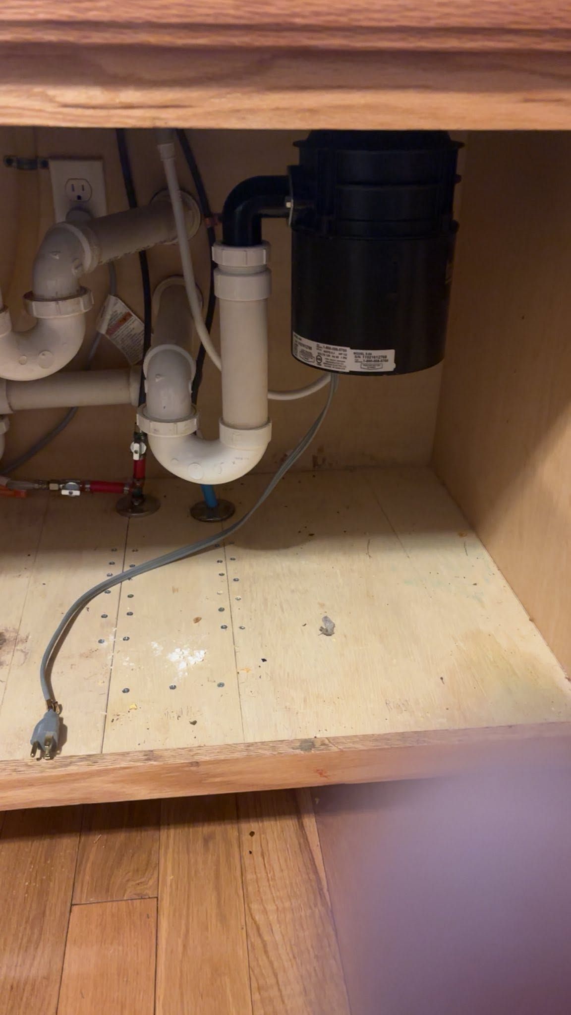 Under-sink plumbing with white pipes, a black disposal, and a wooden cabinet interior with some debris.