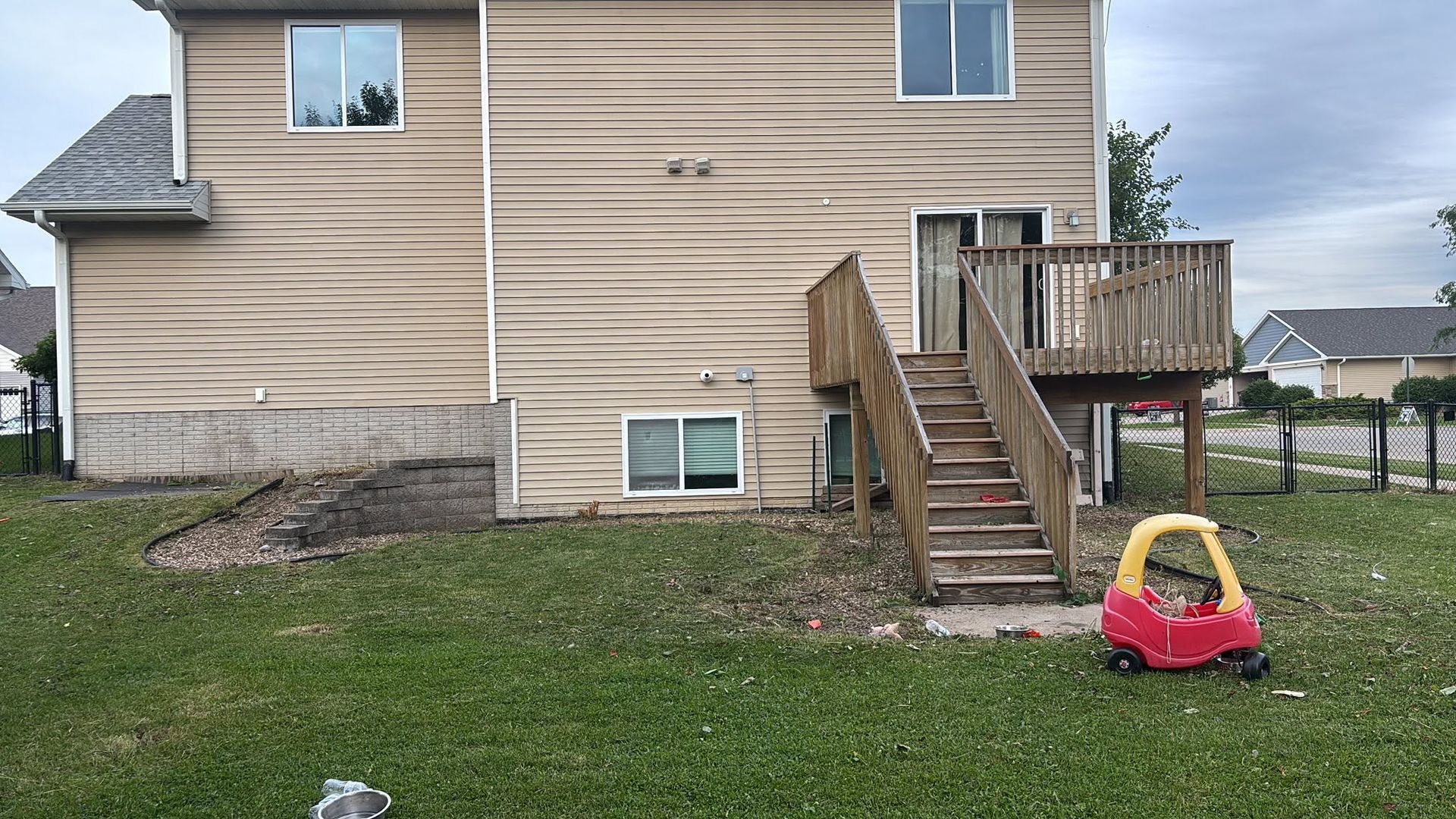 Backyard view of beige house with wooden stairs, deck, and a red-and-yellow toy car on the grass