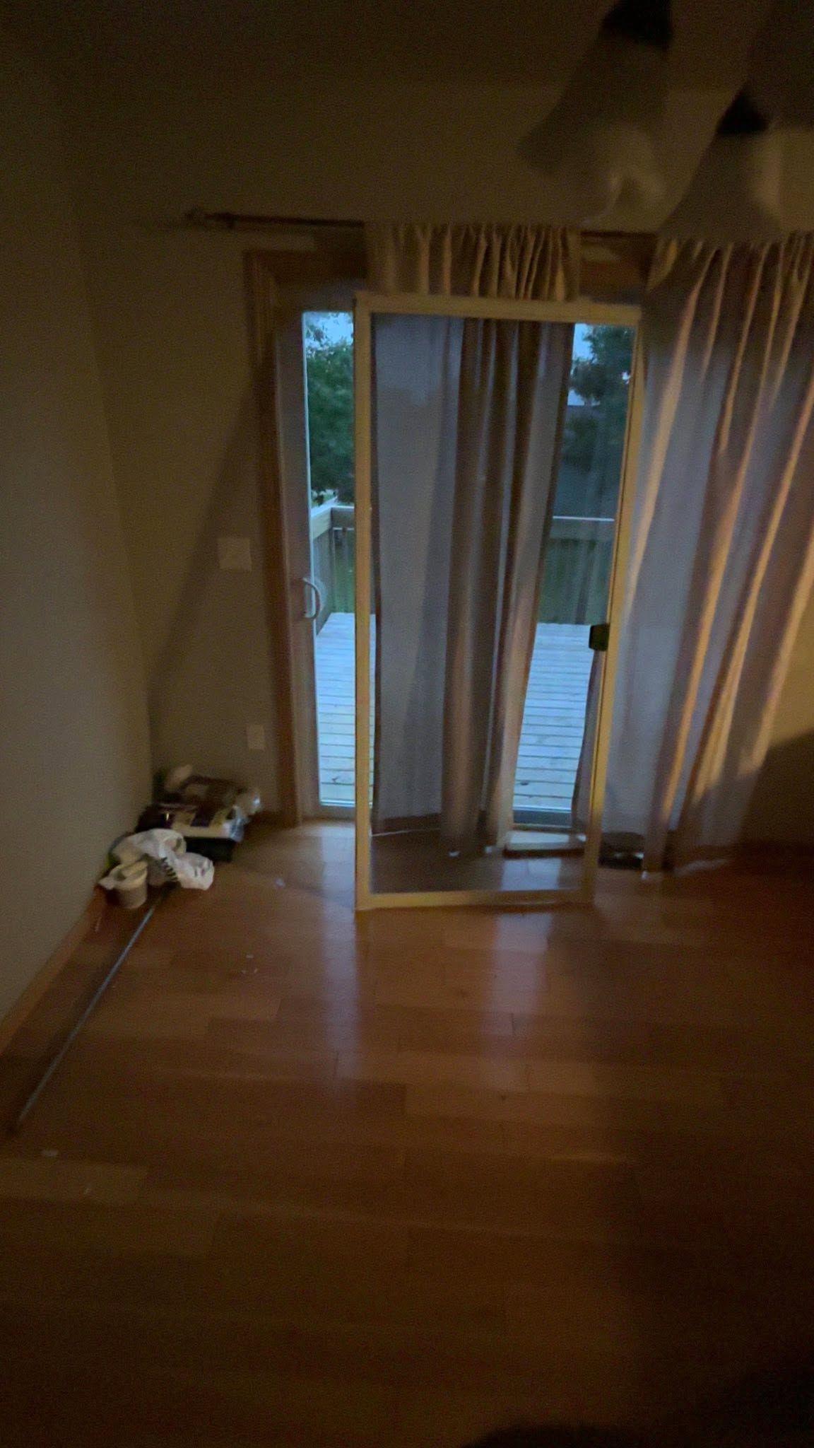 Dim living room with wooden floor, sliding glass doors, and cream curtains overlooking a yard.