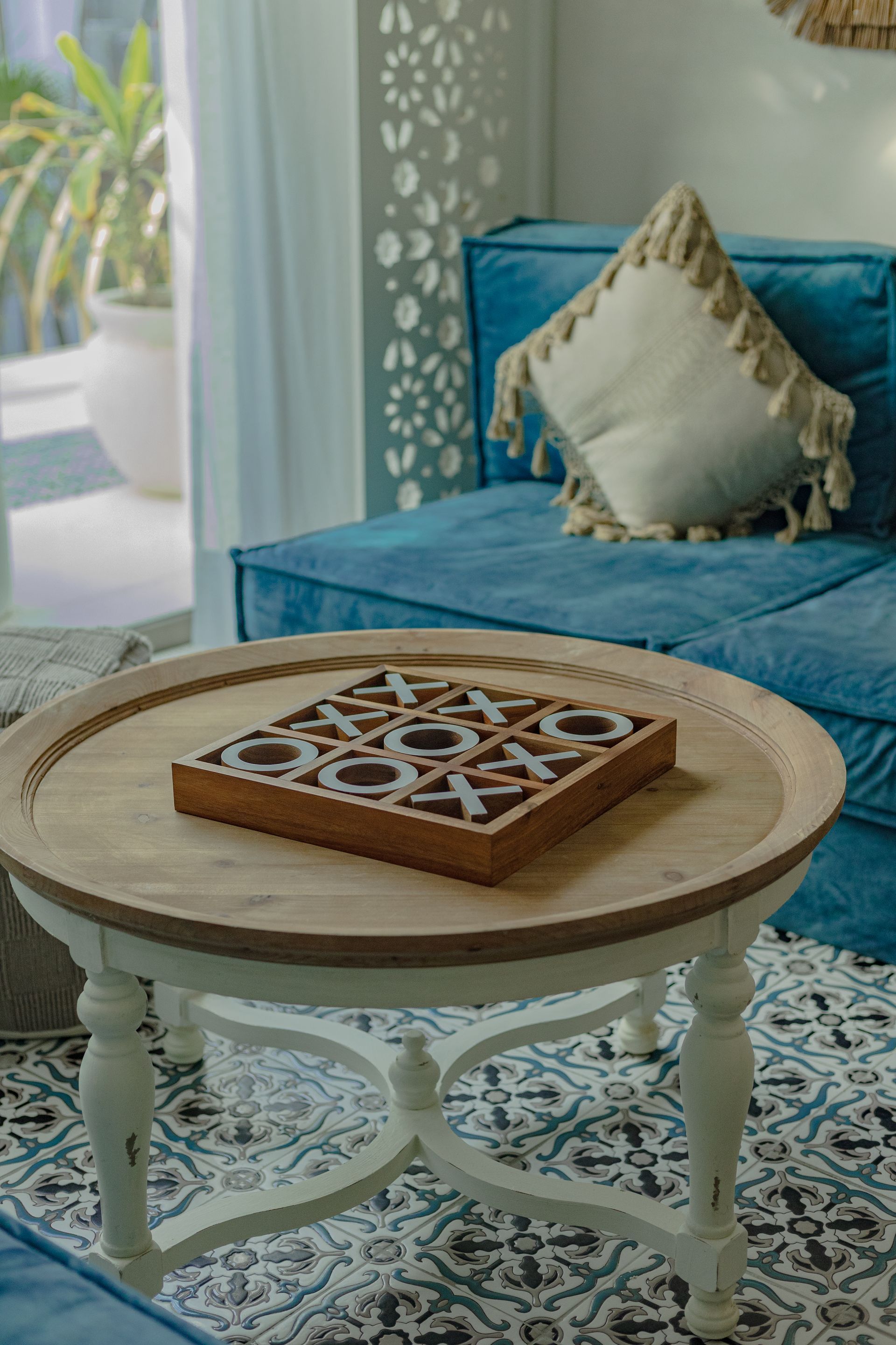 Mesa de centro de madera con tablero de juego, frente a un sofá azul, cerca de una ventana con luz solar.