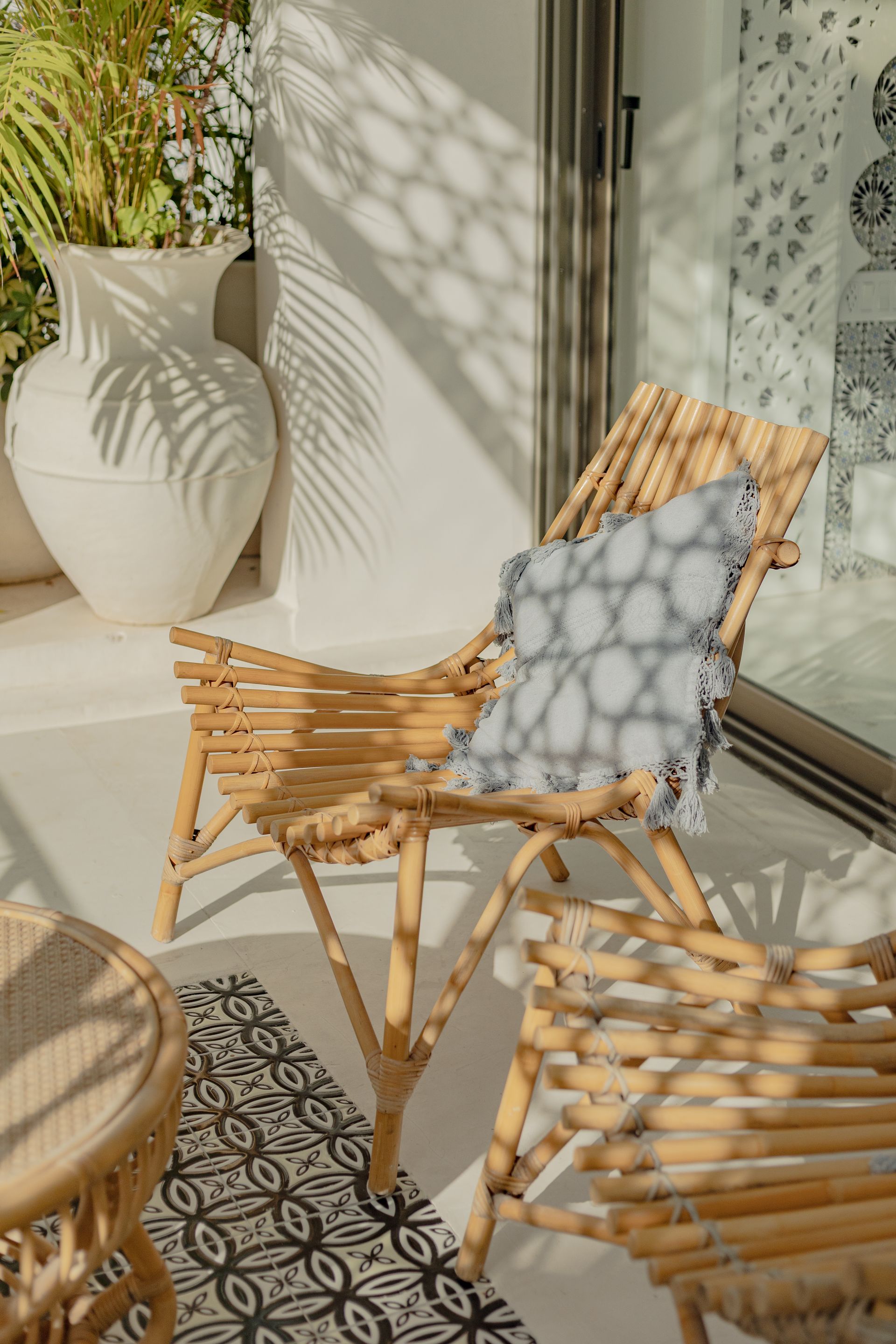 Silla de ratán con cojín en un balcón. Paredes blancas y un jarrón con plantas bajo la luz del sol.