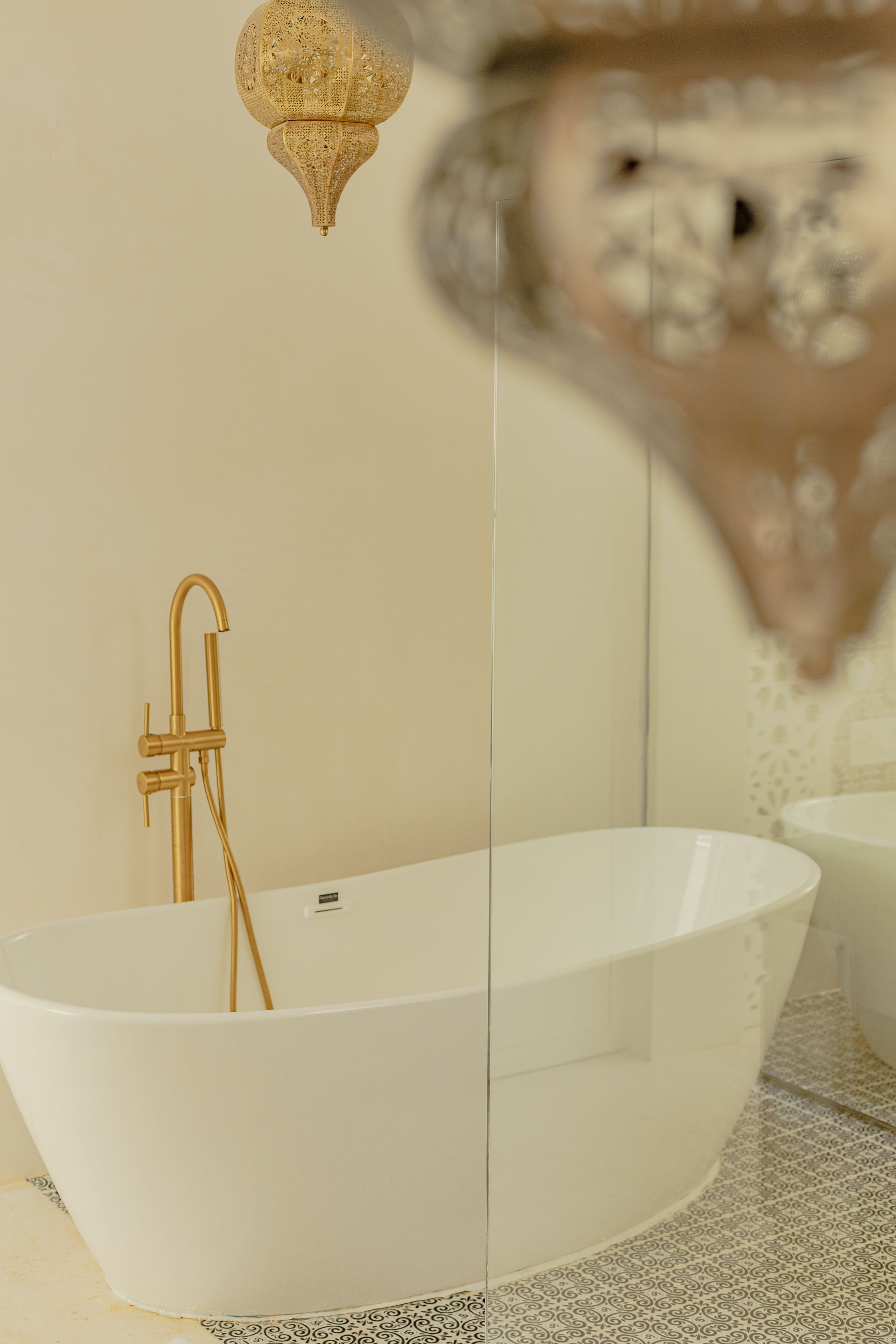 Bañera exenta de color blanco con grifería dorada, en un baño, bajo una luz decorativa dorada.