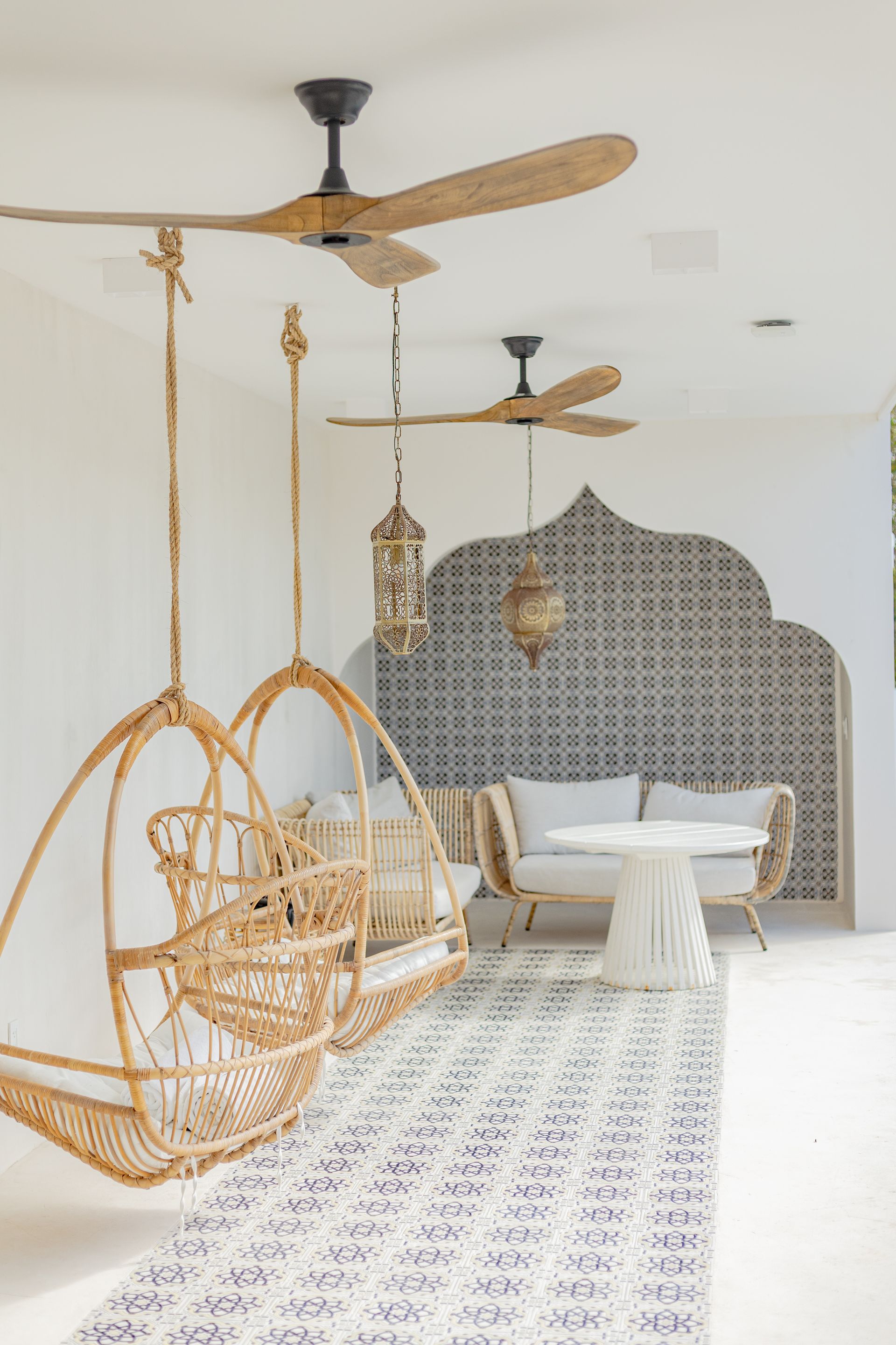 Zona de estar al aire libre con sillas colgantes de mimbre, una mesa pequeña y un patrón de pared decorativo.
