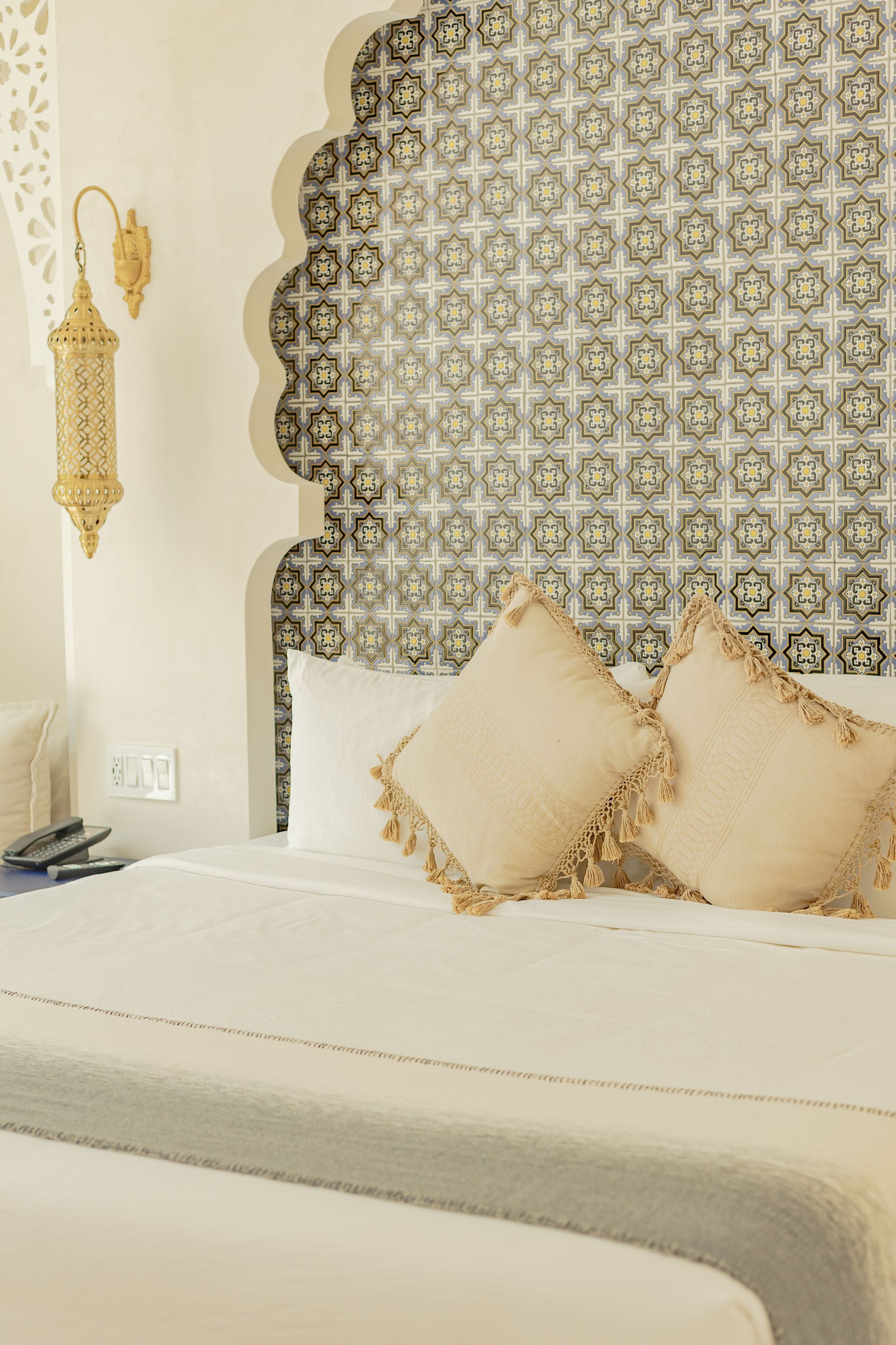 Dormitorio con cabecero de azulejos estampados, ropa de cama blanca y lámpara decorativa dorada.