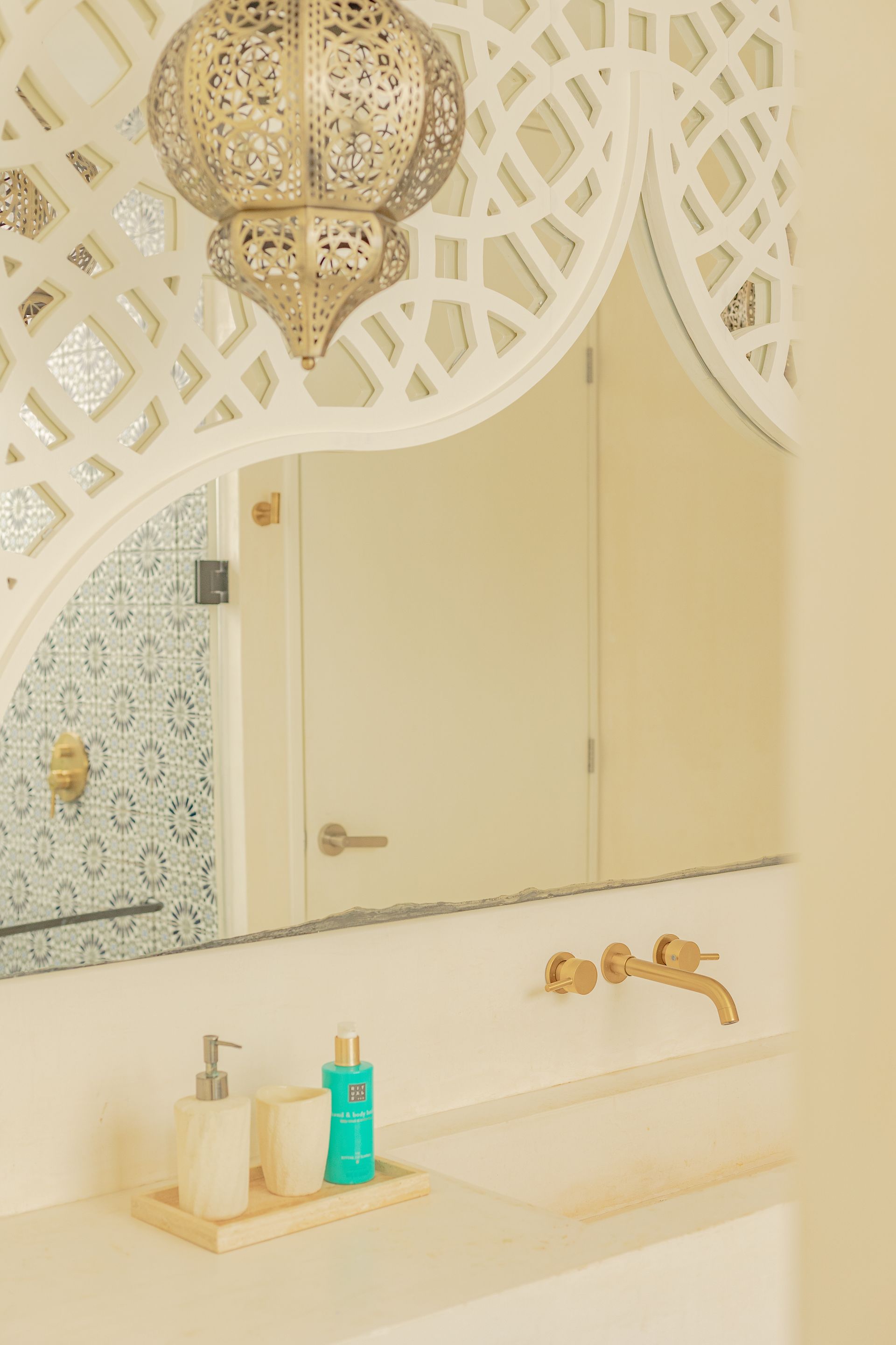 Baño con espejo ornamentado, accesorios de latón y lámpara colgante decorativa. Tonos blanco y dorado.