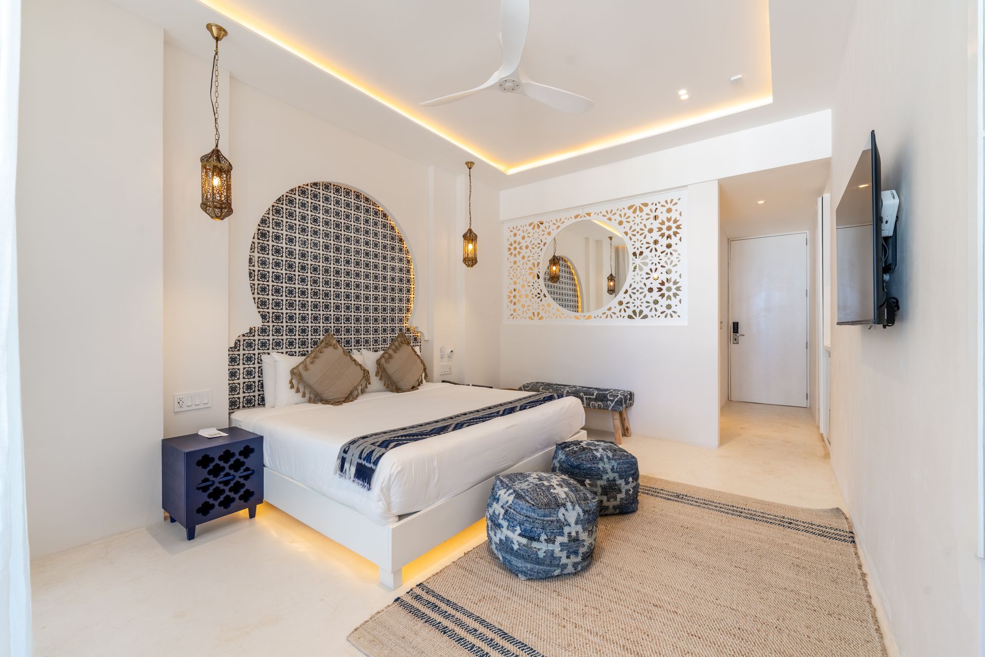 Dormitorio con paredes blancas, cabecero ornamentado y alfombra decorativa. Cuenta con cama, televisor y lámparas colgantes.