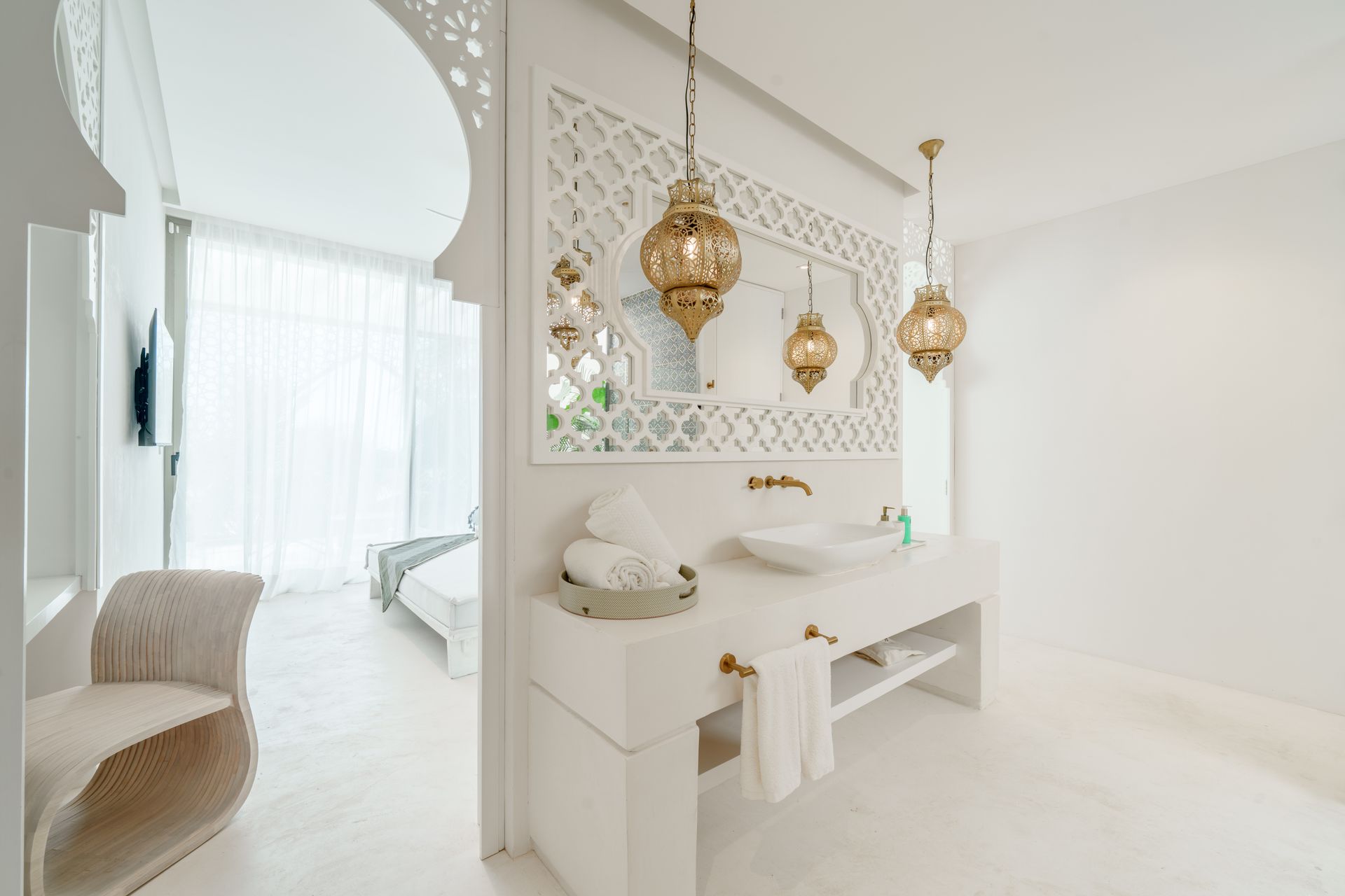 Baño blanco con espejo ornamentado, faroles colgantes y lavabo. Dormitorio visible a través del arco.