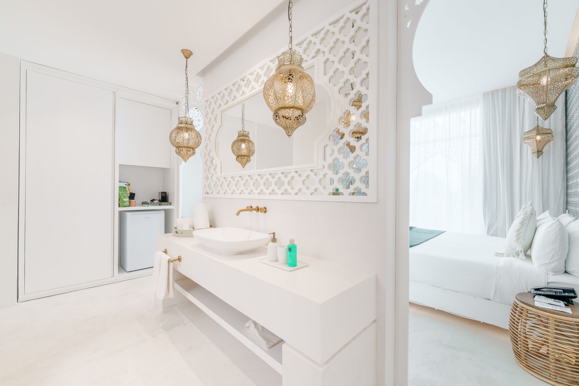 Baño blanco con lámparas colgantes doradas ornamentadas, lavabo y panel decorativo de pared. Dormitorio visible al fondo.