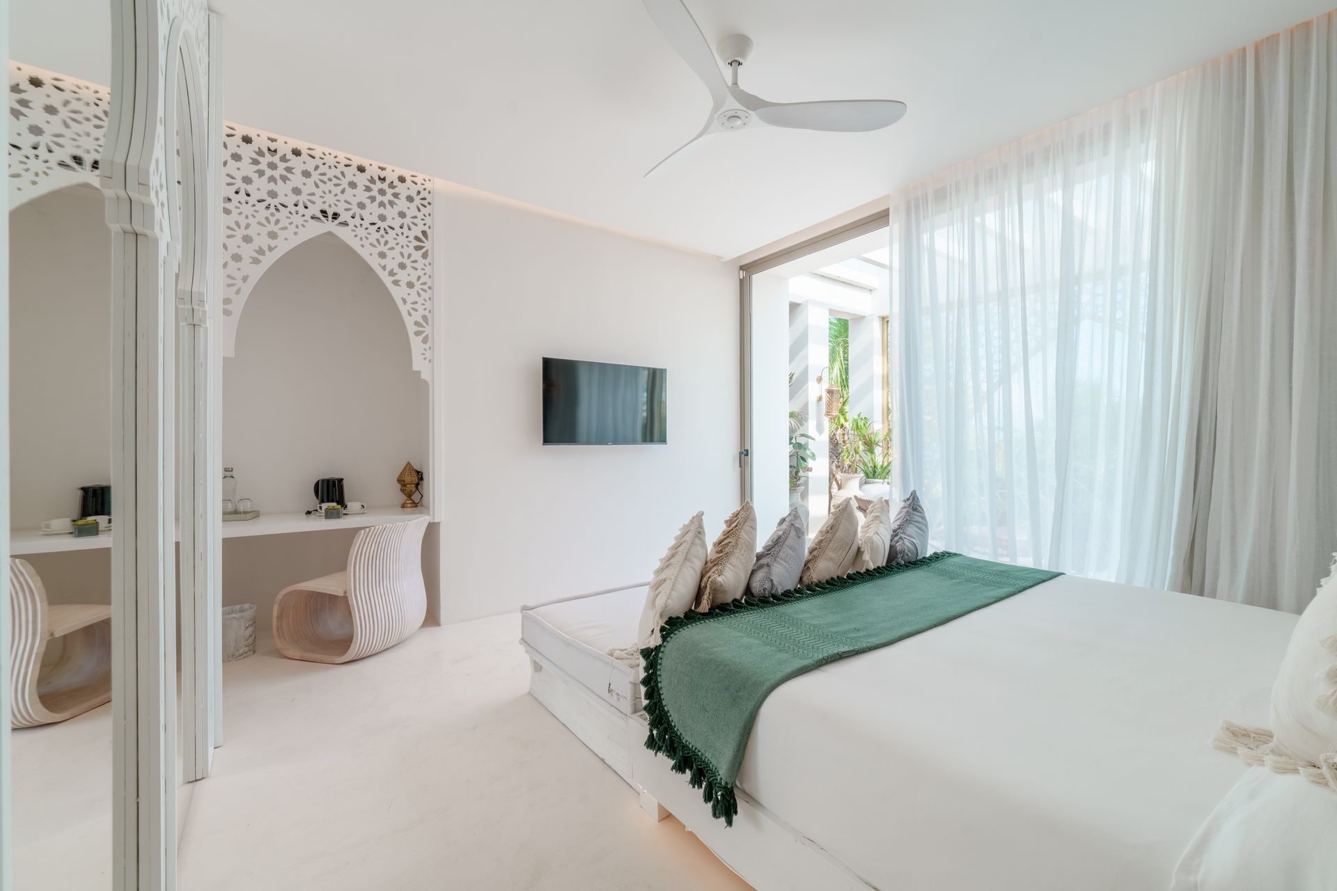 Dormitorio blanco con cabecero ornamentado, escritorio, cama y cortinas transparentes. Una manta verde cubre la cama.