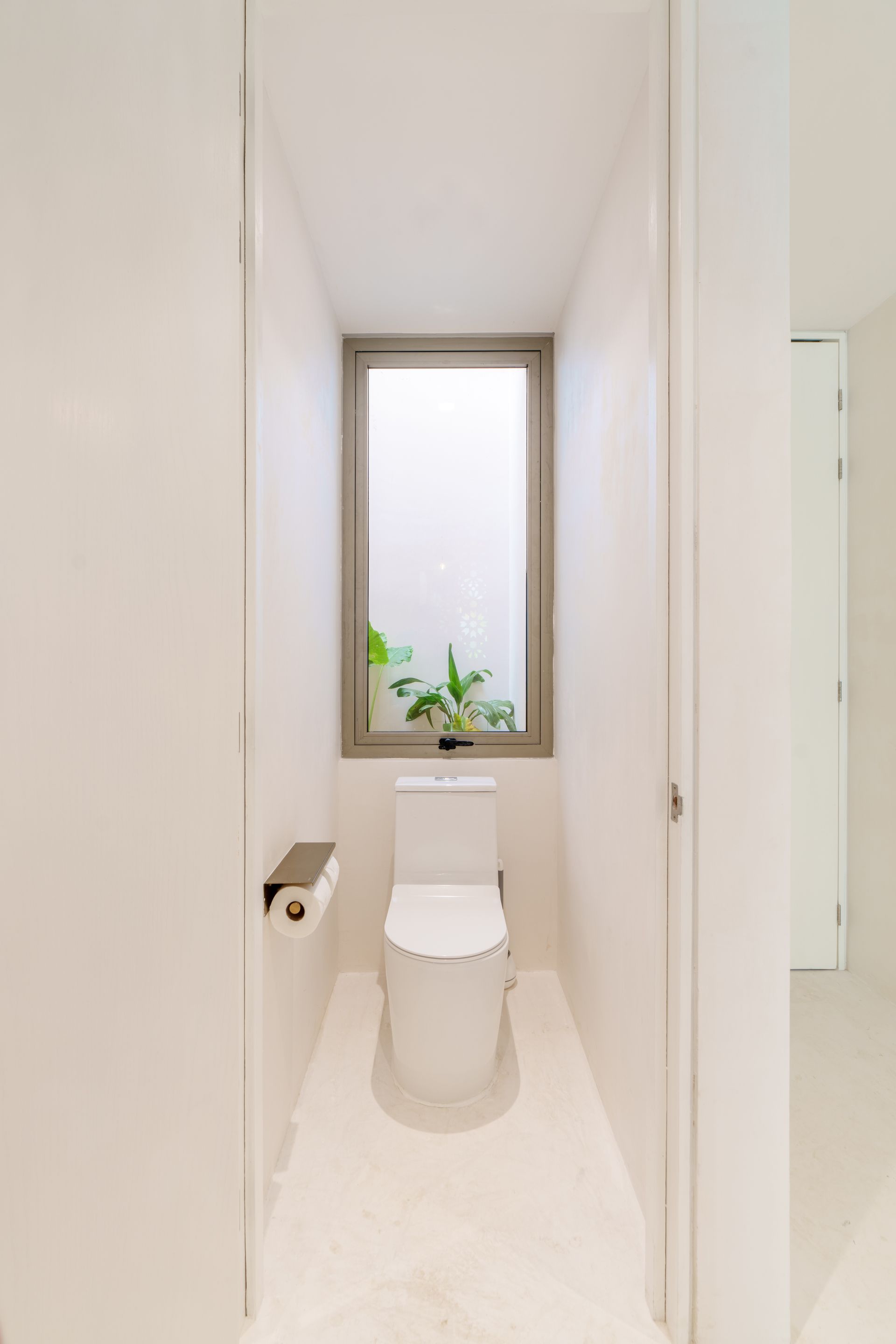 Baño estrecho y blanco con un inodoro centrado debajo de una ventana alta y un soporte para papel higiénico en la pared.