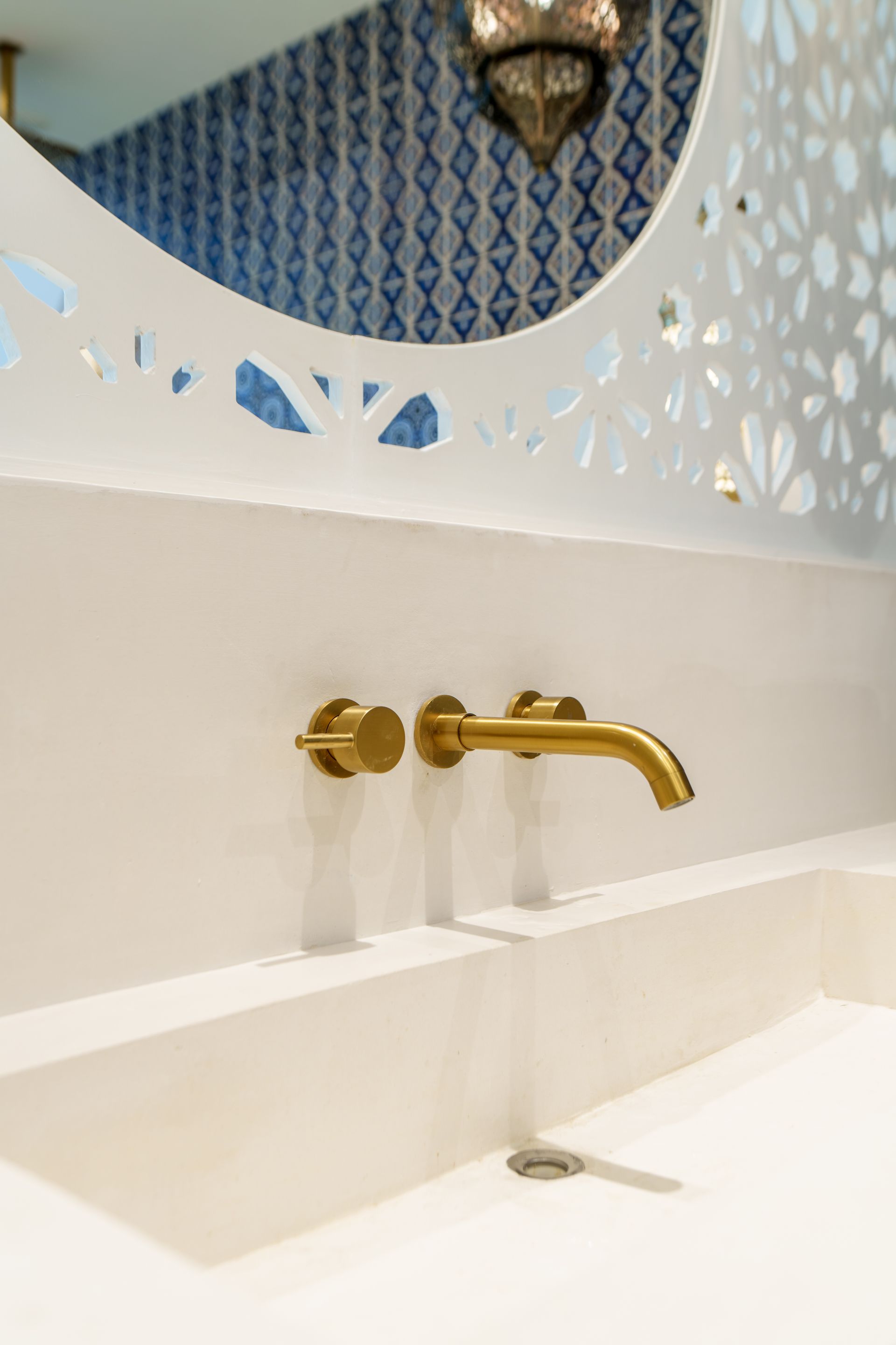Grifo y lavabo dorados sobre pared blanca, bajo un espejo decorativo con fondo de azulejos azules y blancos.