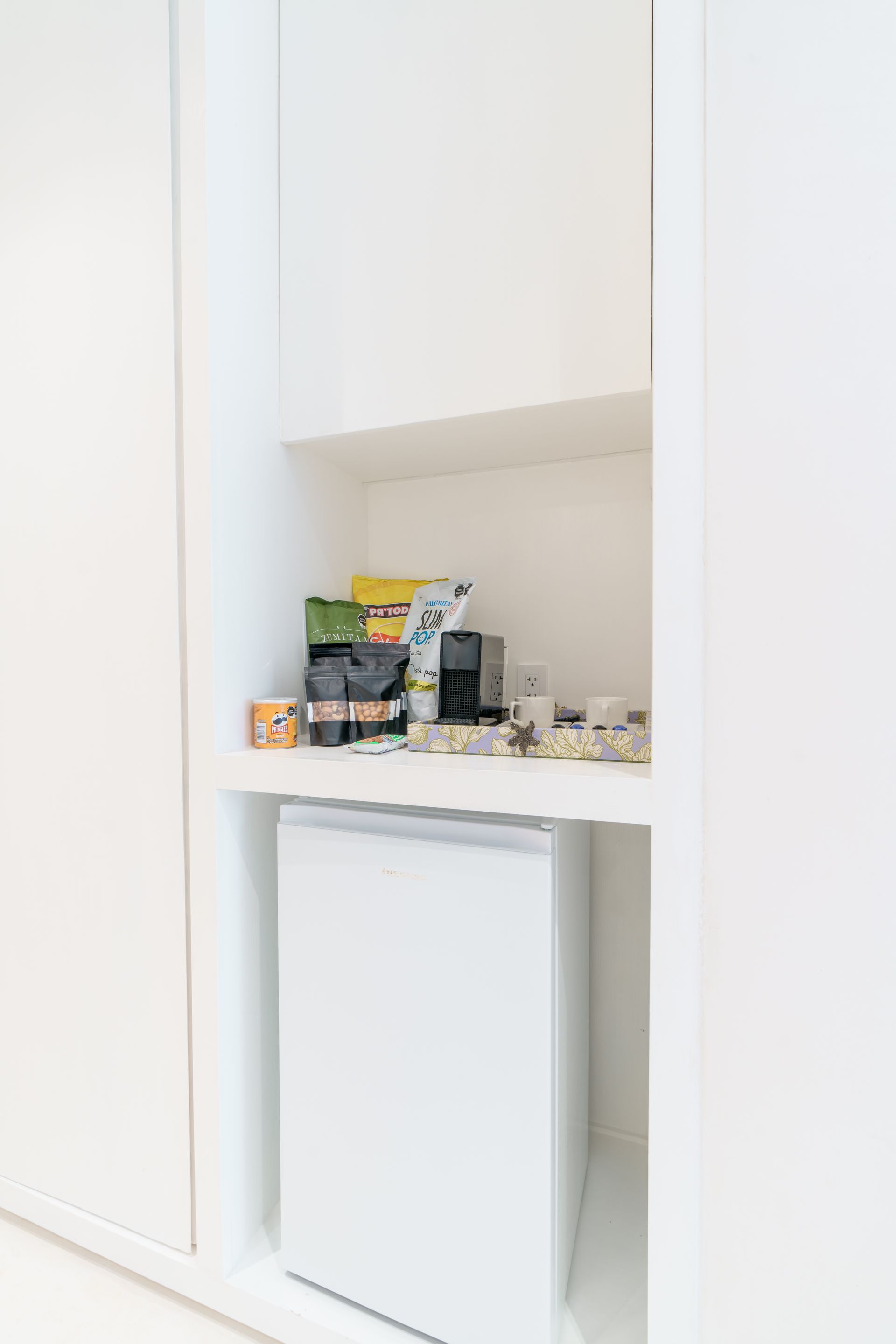 Mueble empotrado de color blanco con mininevera debajo de un estante con snacks y cafetera.
