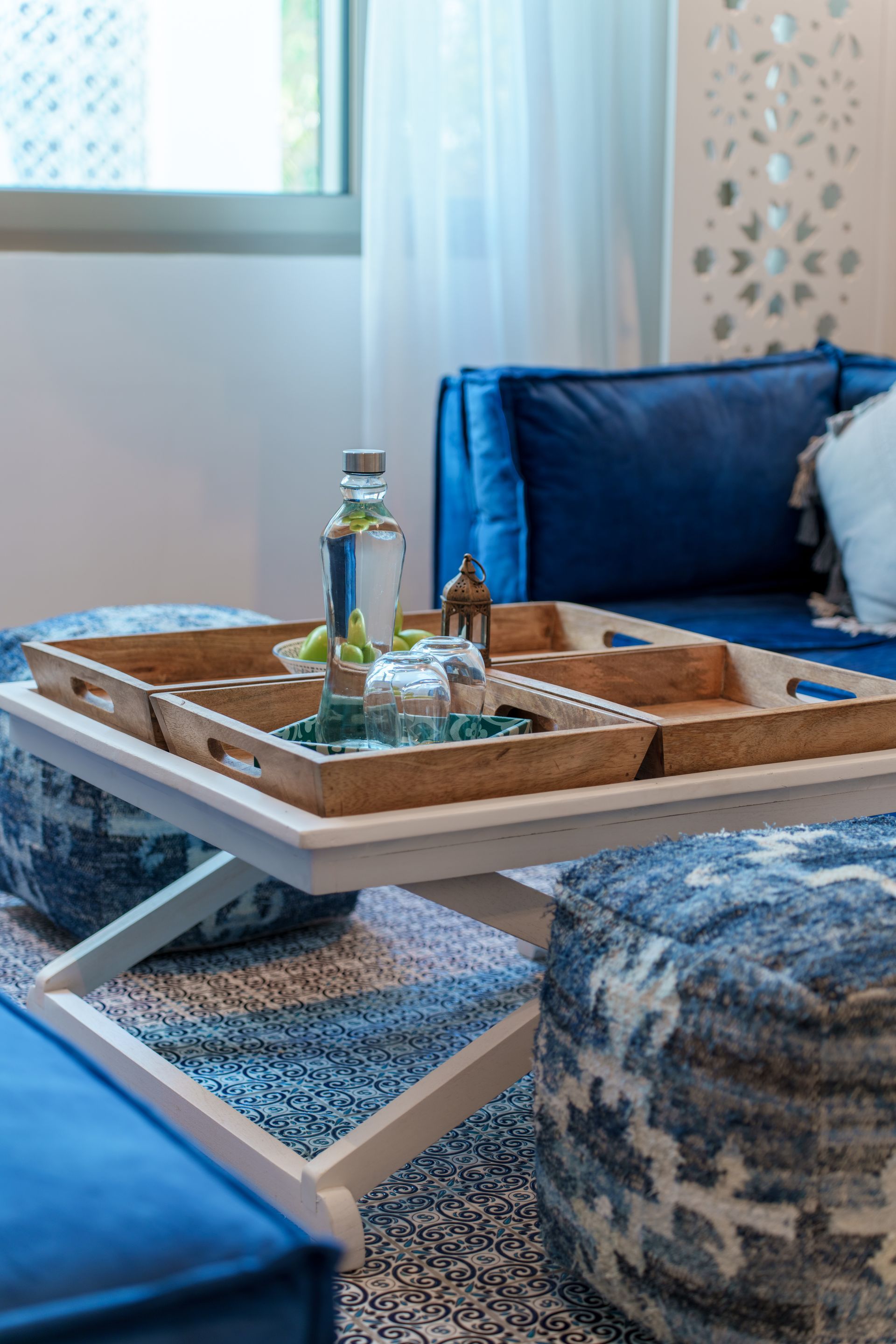 Sala de estar azul con asientos otomanos, una bandeja de madera con bebidas sobre una mesa blanca y una alfombra estampada.