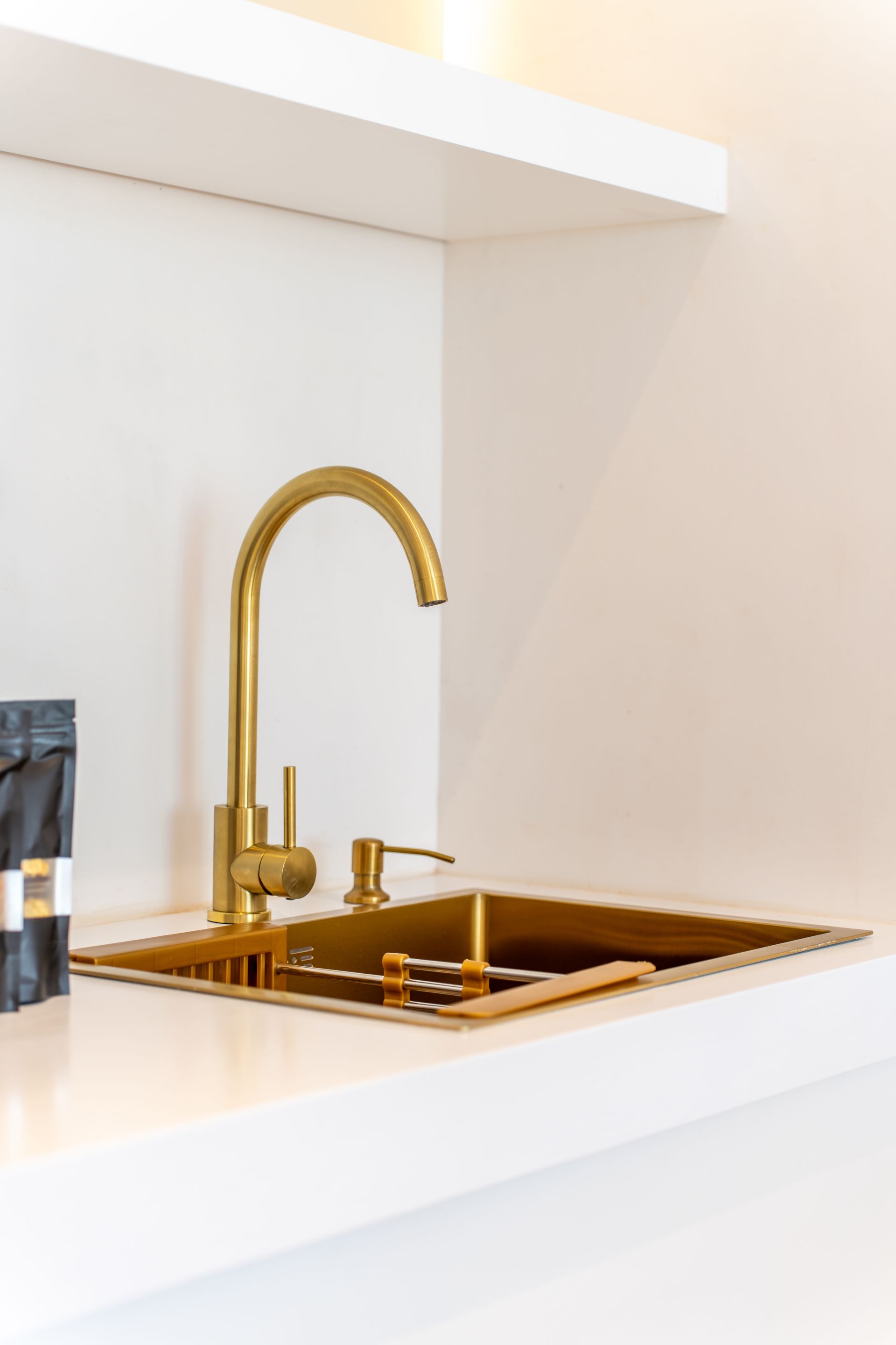 Grifo de cocina dorado y fregadero sobre una encimera y una pared blancas, con un estante encima.