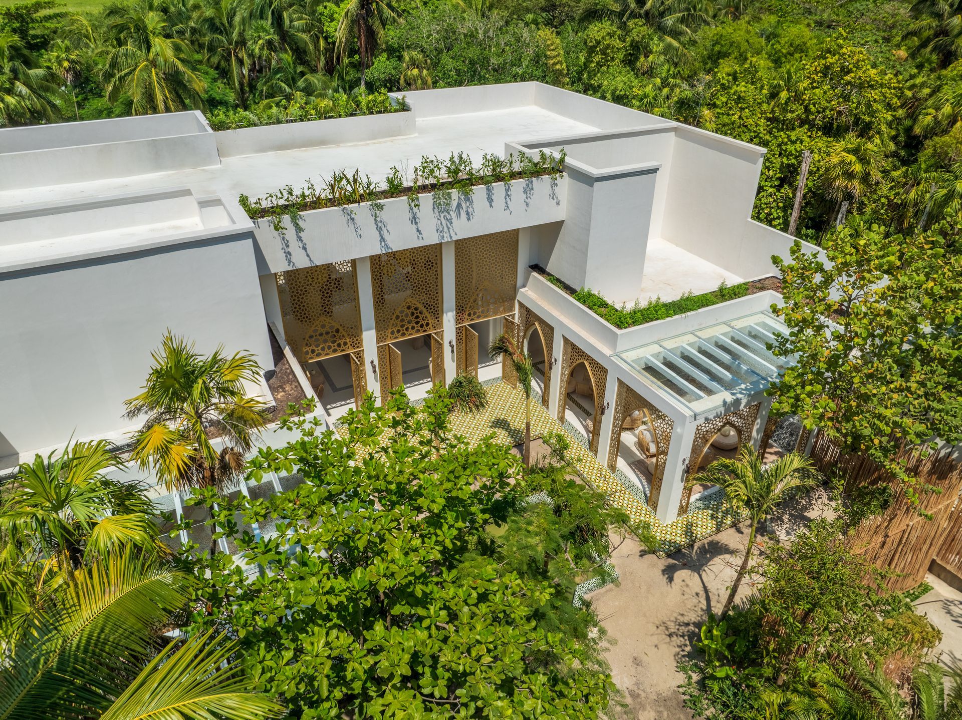 Edificio blanco rodeado de exuberantes árboles verdes, con detalles arquitectónicos que incluyen plantas colgantes y una pérgola.