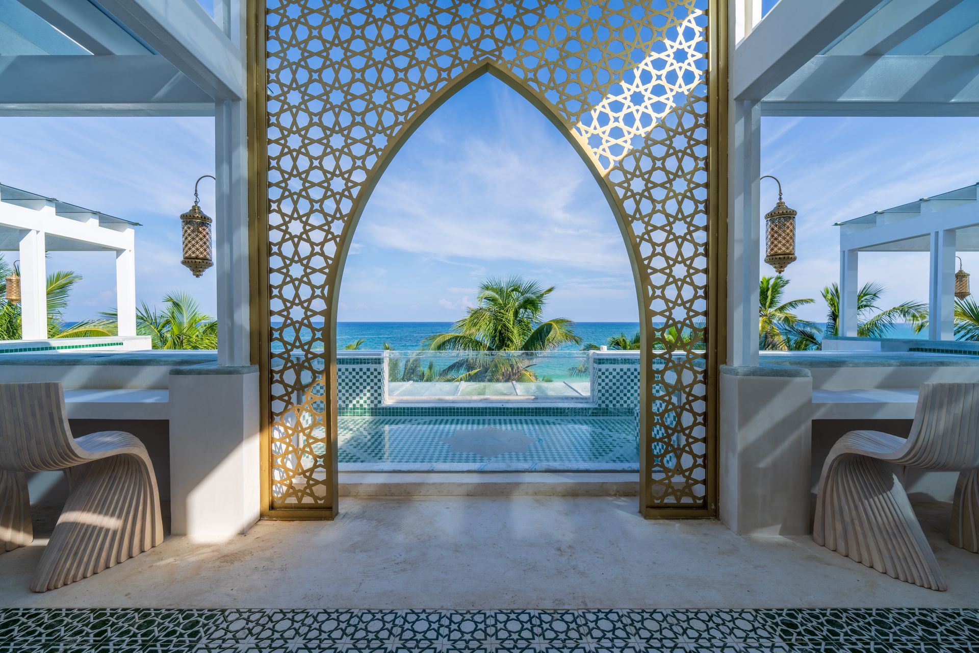 Un arco dorado ornamentado enmarca la vista al mar; pabellón blanco con linternas colgantes y piso estampado.