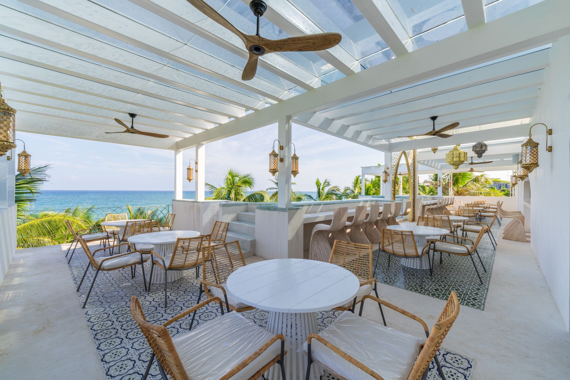 Bar al aire libre con vista al mar, decoración blanca, sillas de mimbre, ventiladores de techo e iluminación cenital.