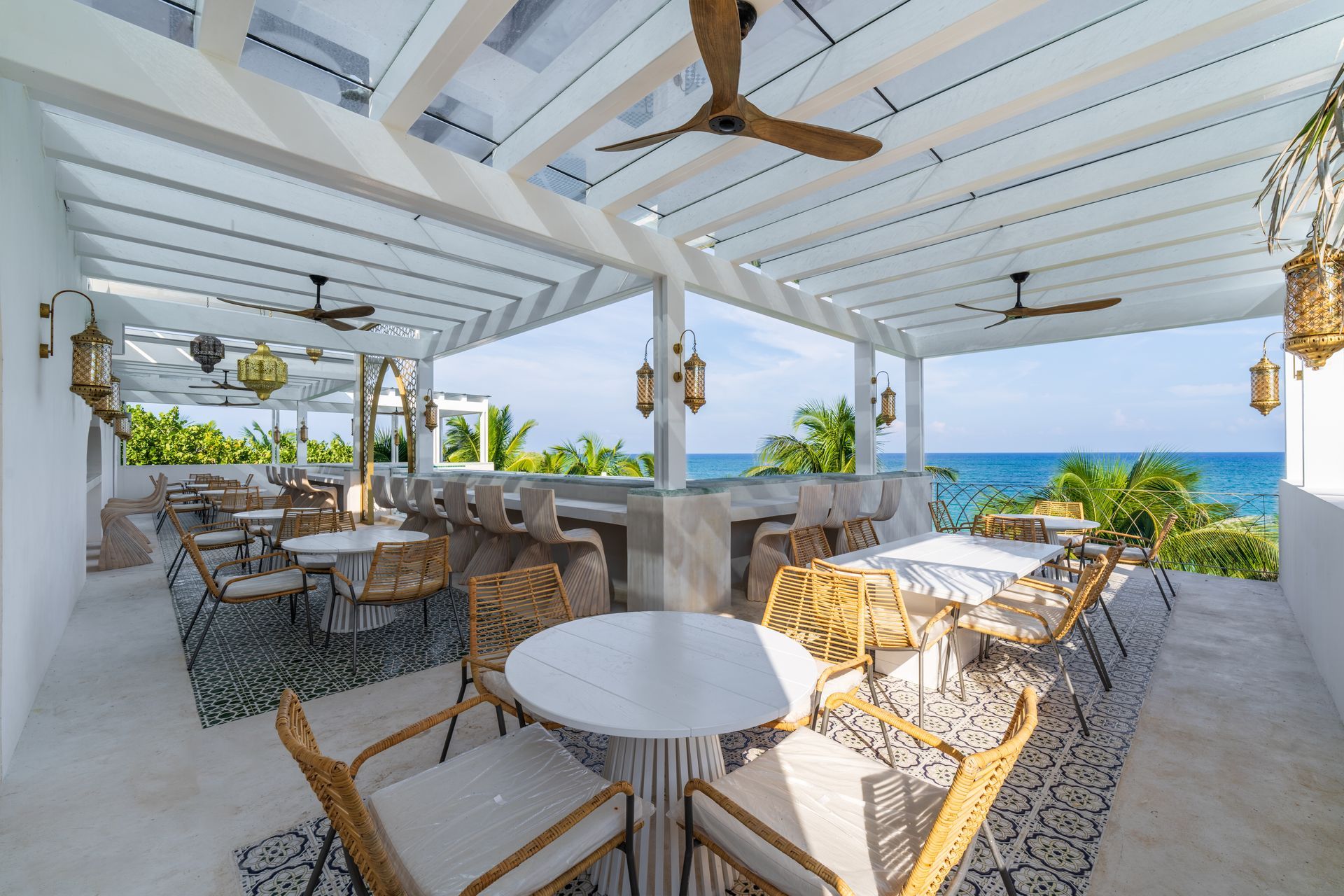 Restaurante al aire libre con mesas, sillas, bar y vista al mar bajo pérgola blanca.