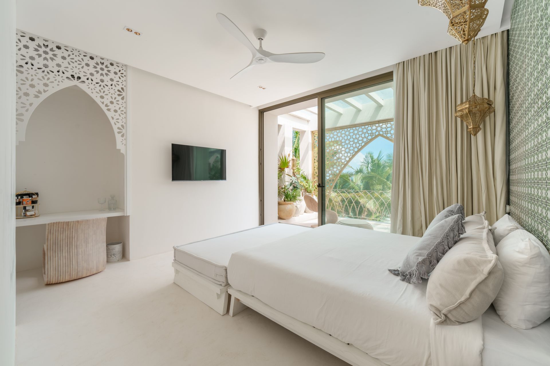 Dormitorio blanco con cama, TV, decoración de pared ornamentada y puerta corrediza de vidrio a un balcón.