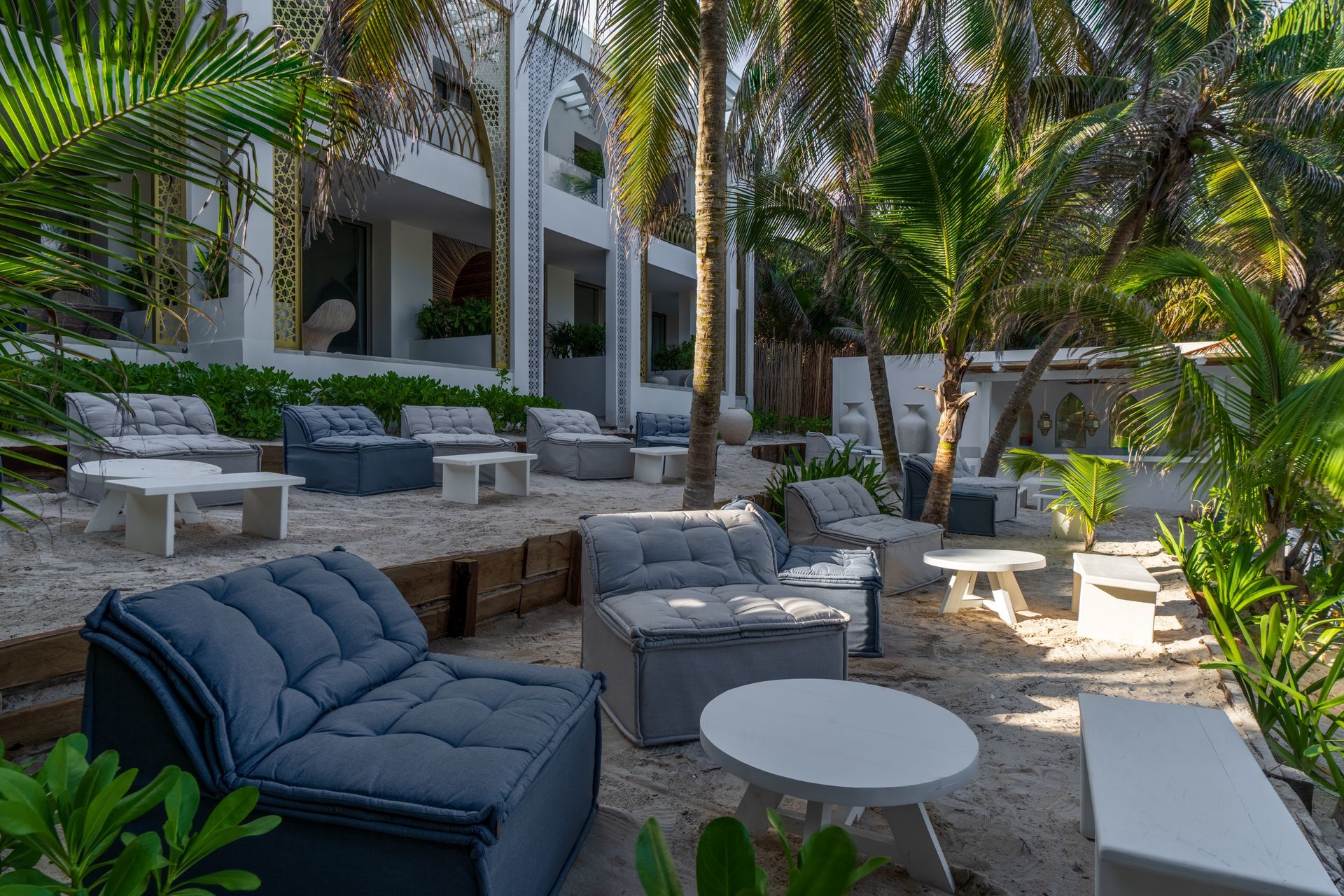Zona de salón al aire libre con mesas blancas, cojines azules y palmeras.