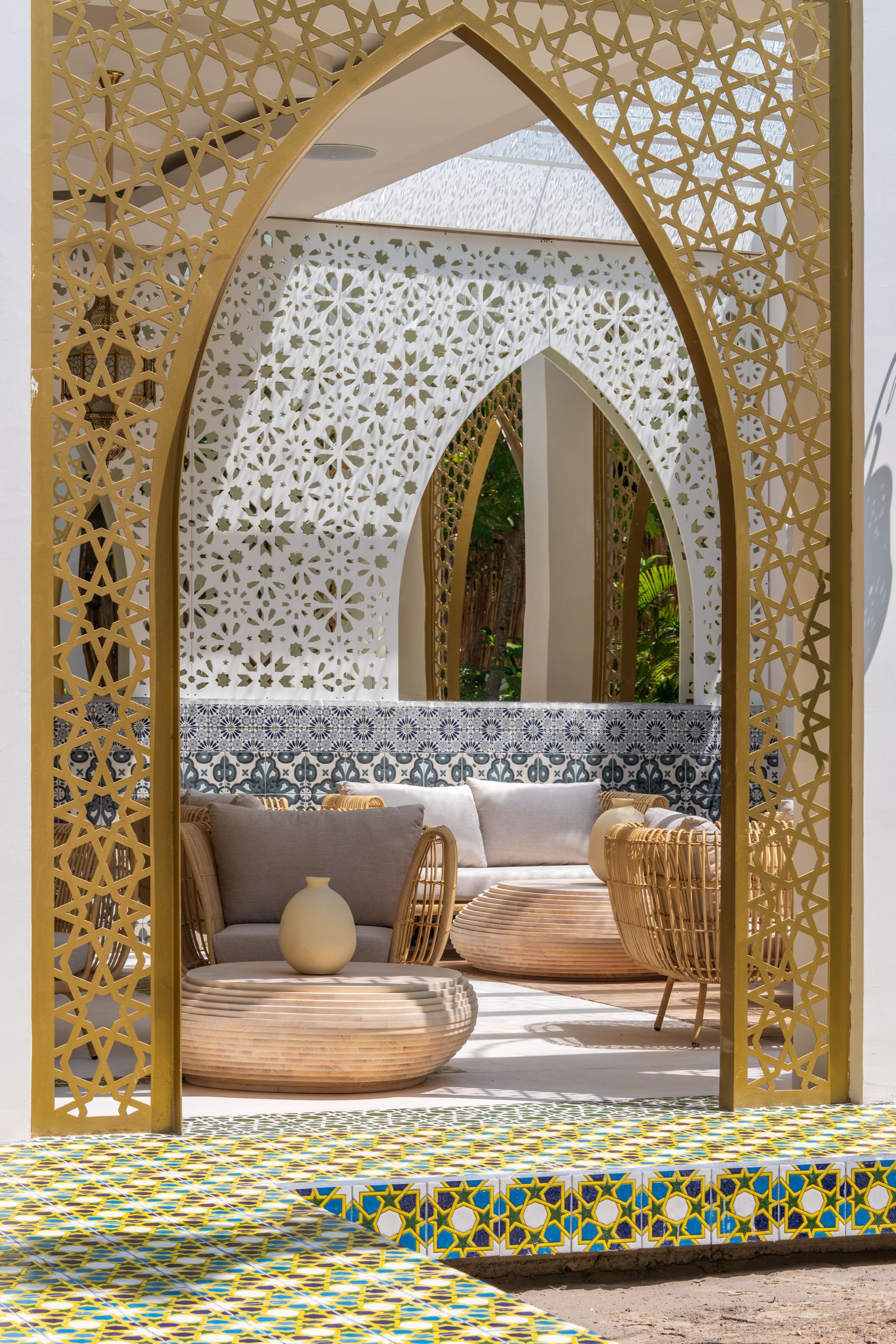 Un arco dorado ornamentado enmarca el área de asientos al aire libre: muebles de ratán, almohadas blancas, piso de baldosas estampadas y detalles de paredes recortadas.
