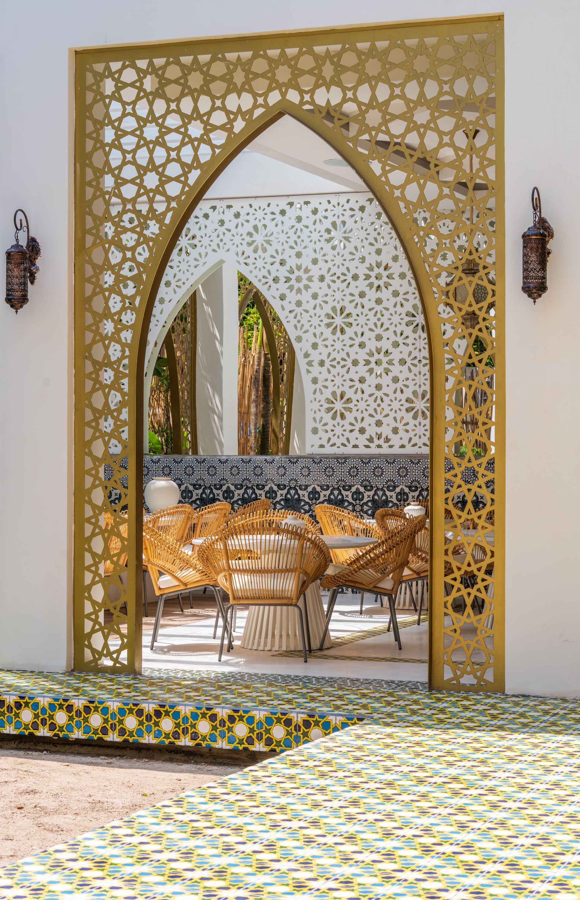 Puerta dorada adornada, con recortes estampados, enmarca el salón interior con muebles de mimbre, paredes blancas y azulejos decorativos.