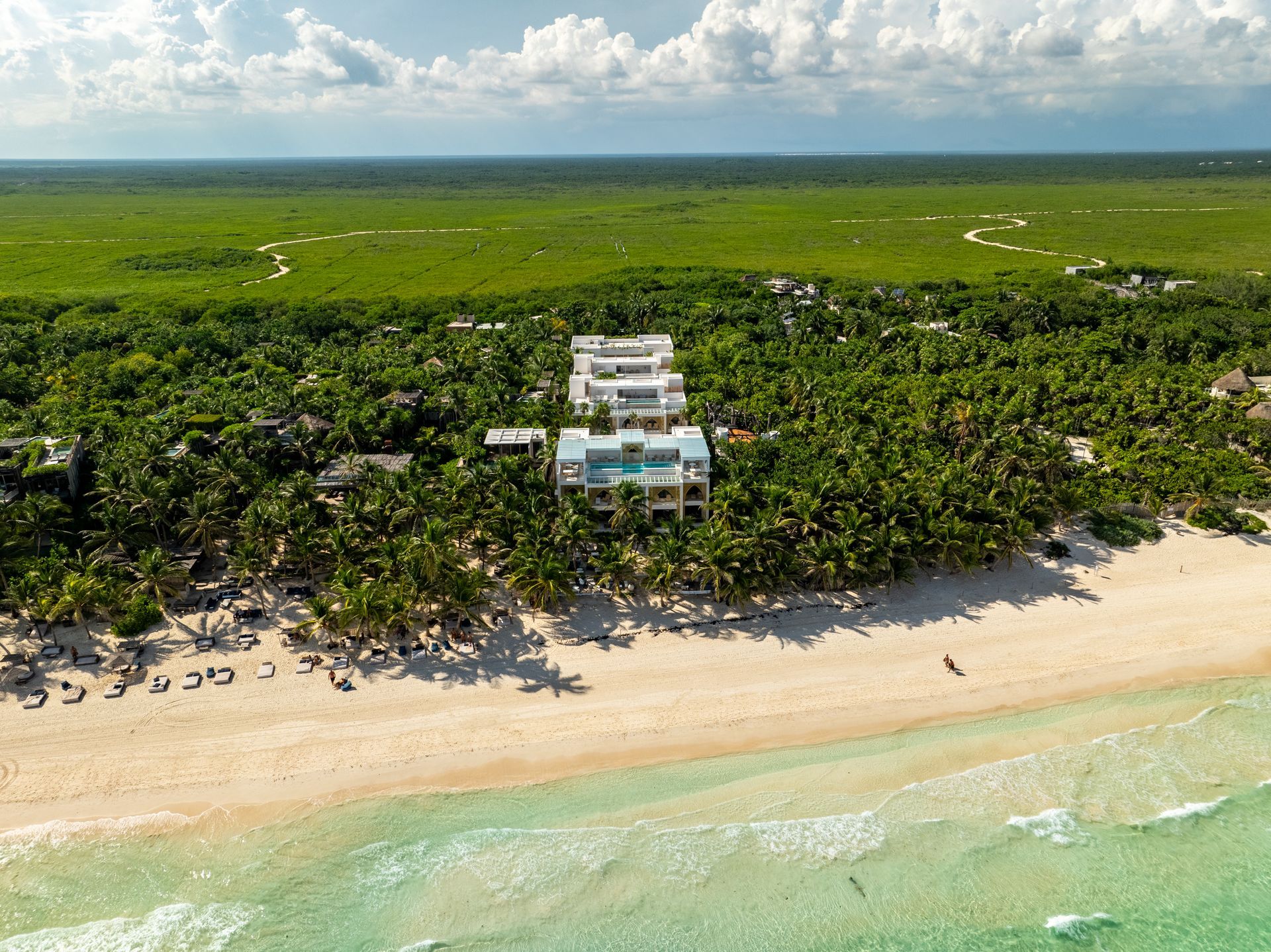 Vista aérea de villas frente a la playa rodeadas de exuberantes árboles verdes y una playa de arena blanca con agua turquesa.