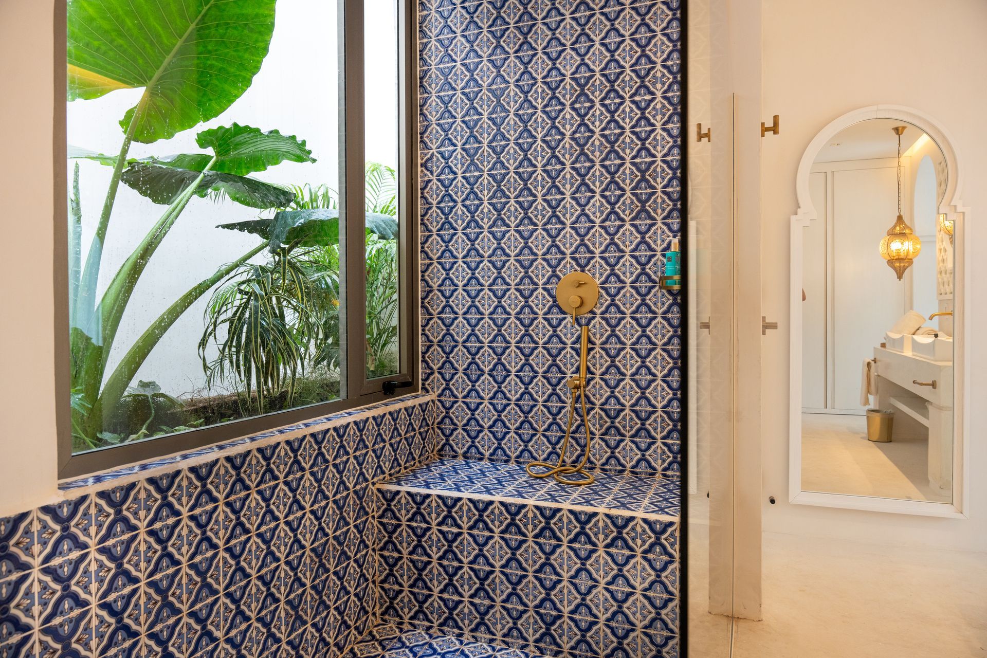 Baño con ducha con azulejos con estampados azules y blancos, ventana con follaje verde y tocador blanco con un espejo adornado.