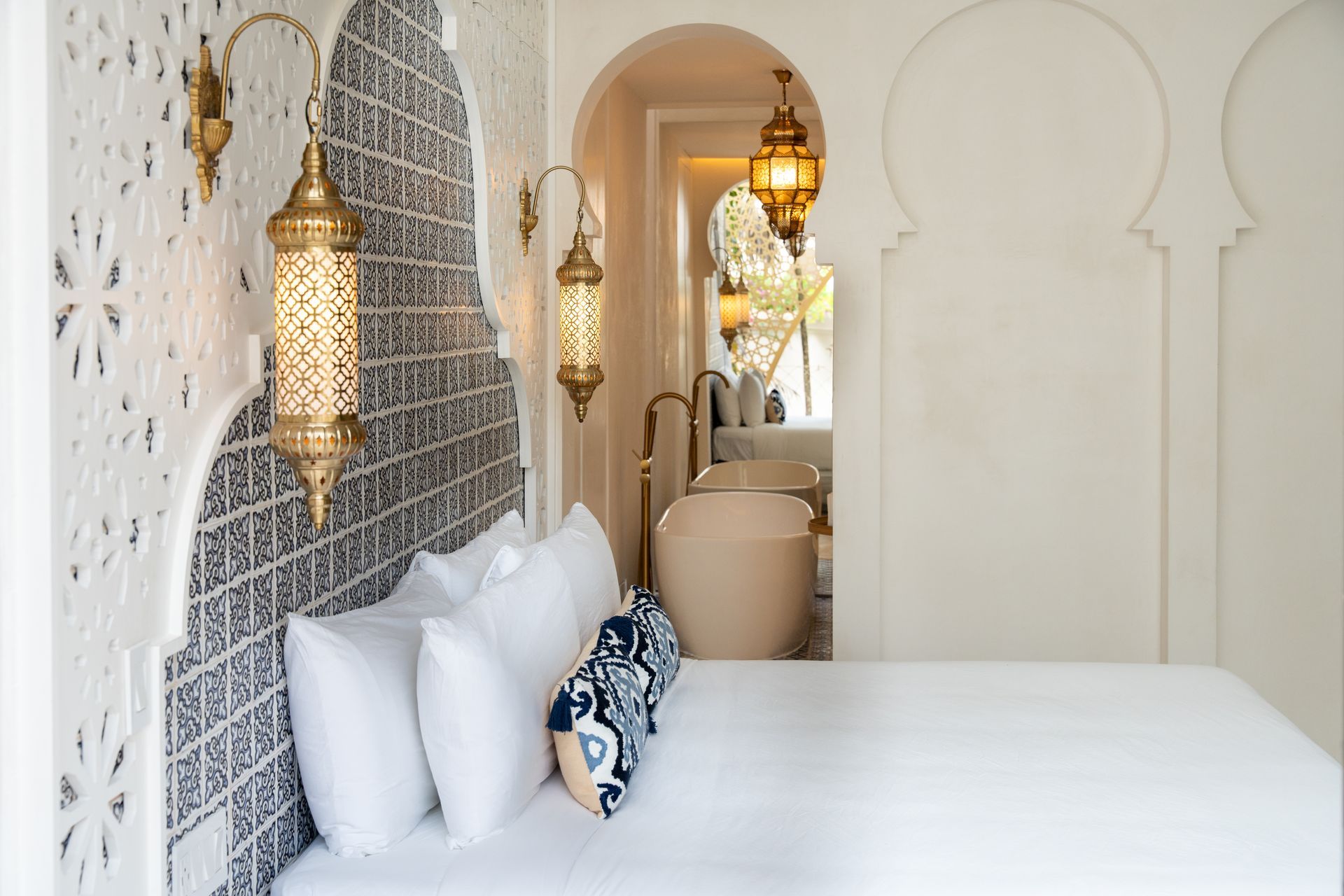 Dormitorio con decoración de estilo marroquí: lámparas ornamentadas, puertas arqueadas, pared con detalles estampados y vista a un baño.