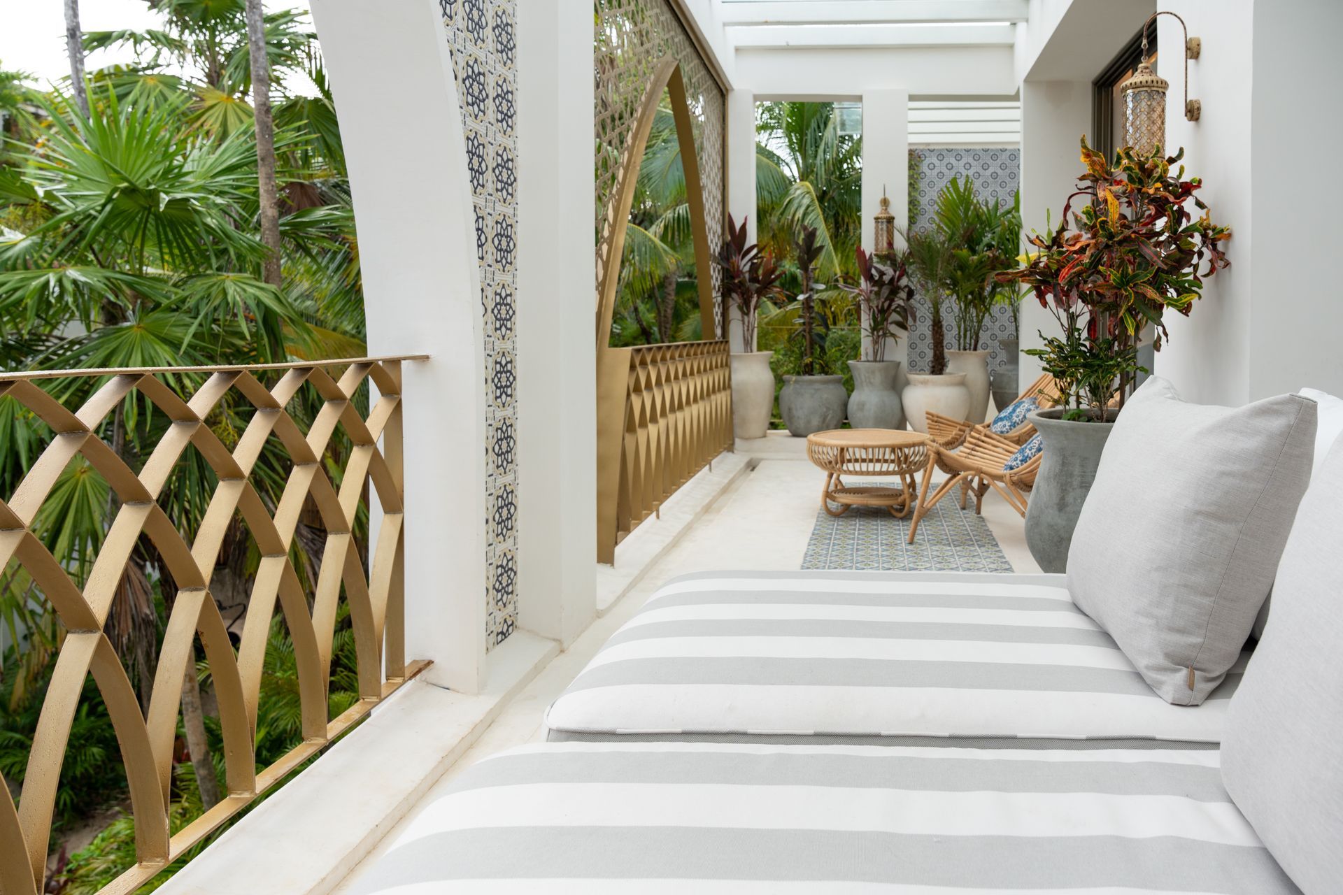 Balcón con salón de rayas, barandillas doradas, plantas en macetas y follaje tropical.