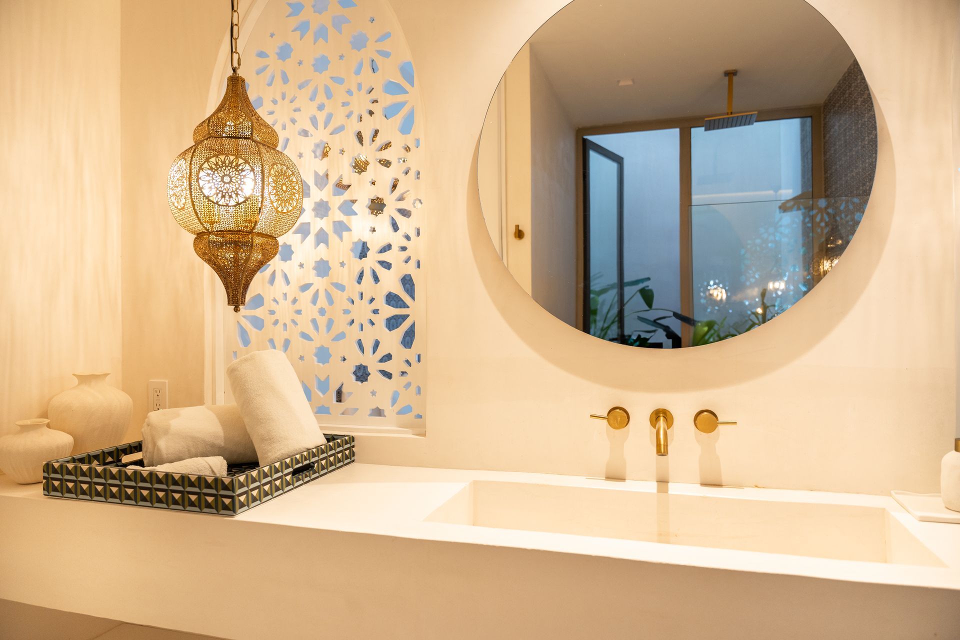 Baño de estilo marroquí con accesorios dorados, una linterna decorativa y un espejo redondo que refleja una vista exterior.