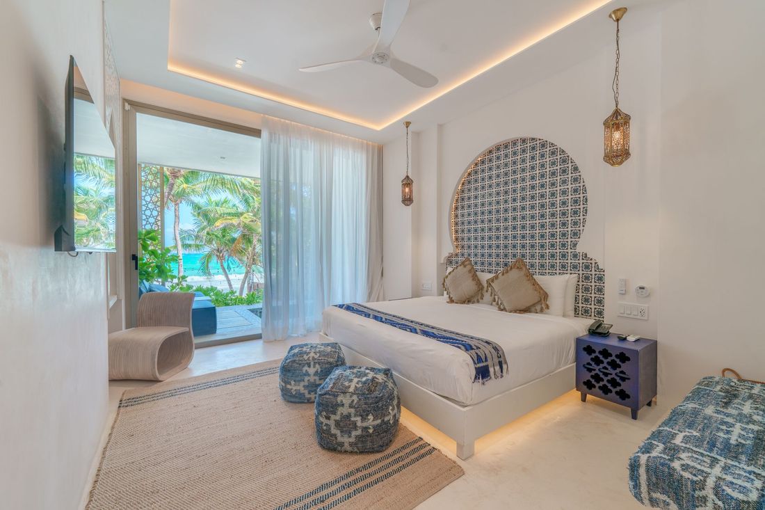 Bright bedroom with beach view, white walls, ornate headboard, blue accents, sliding glass doors.