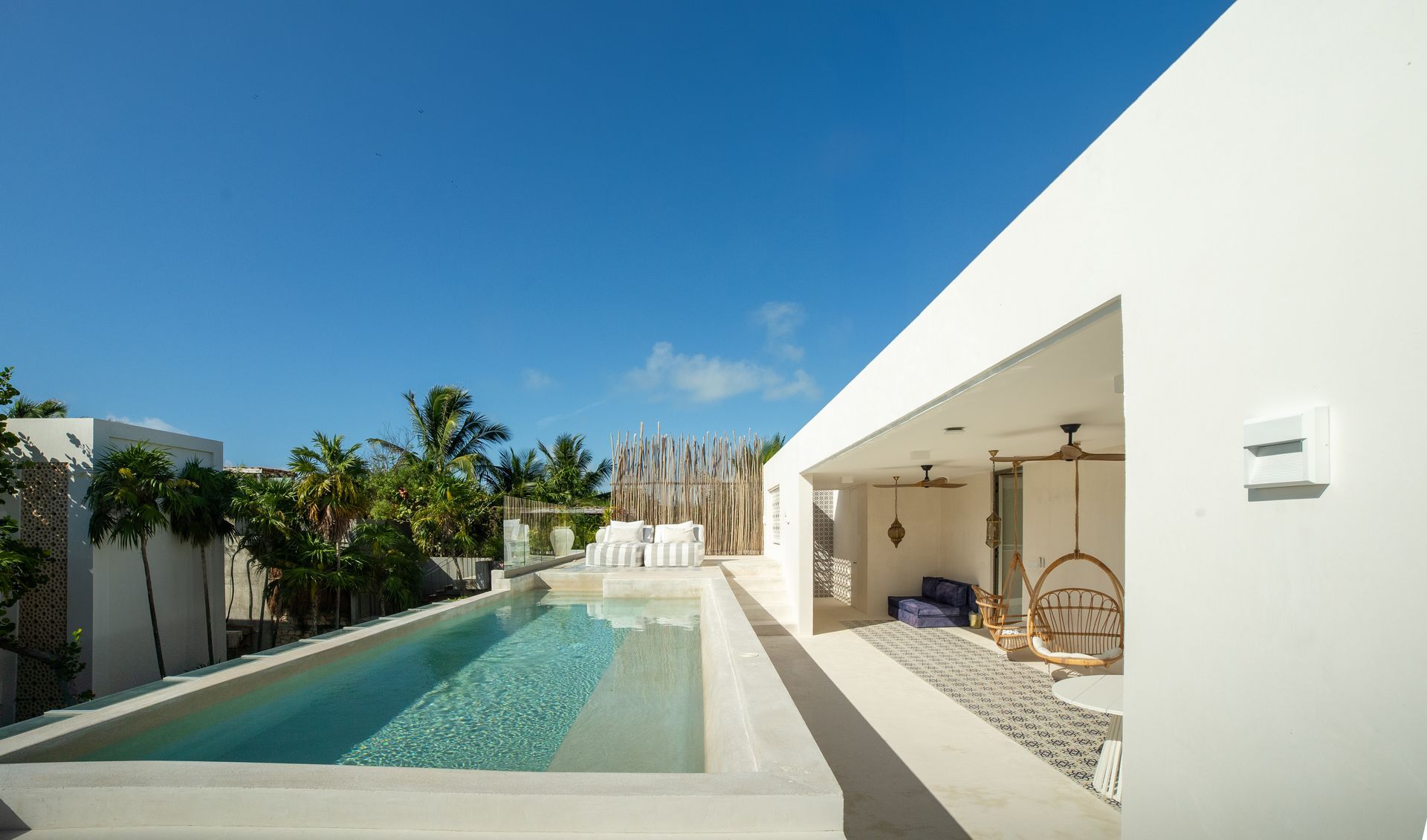 Piscina en la azotea y patio a la sombra en un día soleado. Paredes blancas, cielo azul y vegetación tropical.