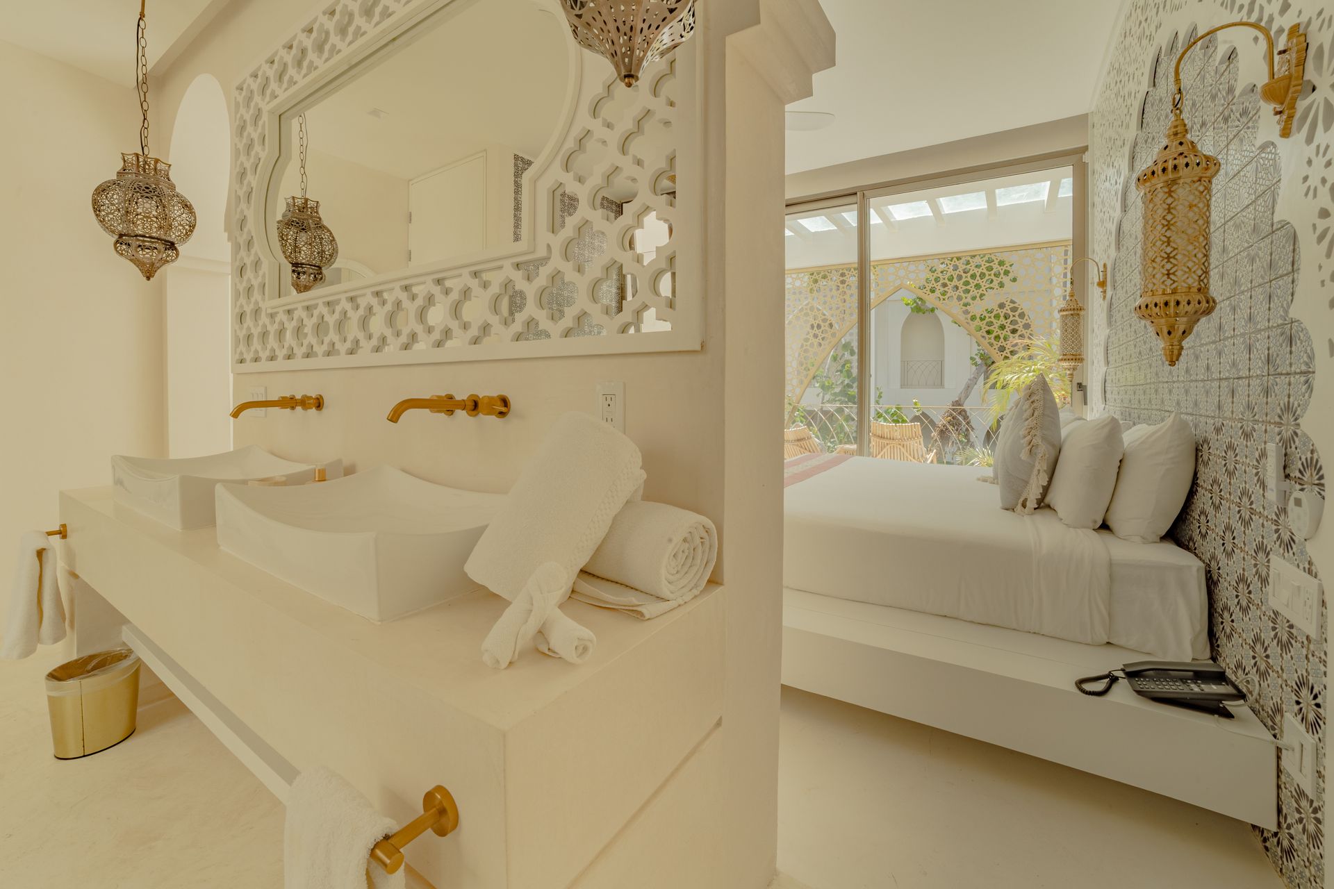 Baño con espejo ornamentado  y vista a una cama a través de una abertura. Decoración  en blancos con detalles dorados.