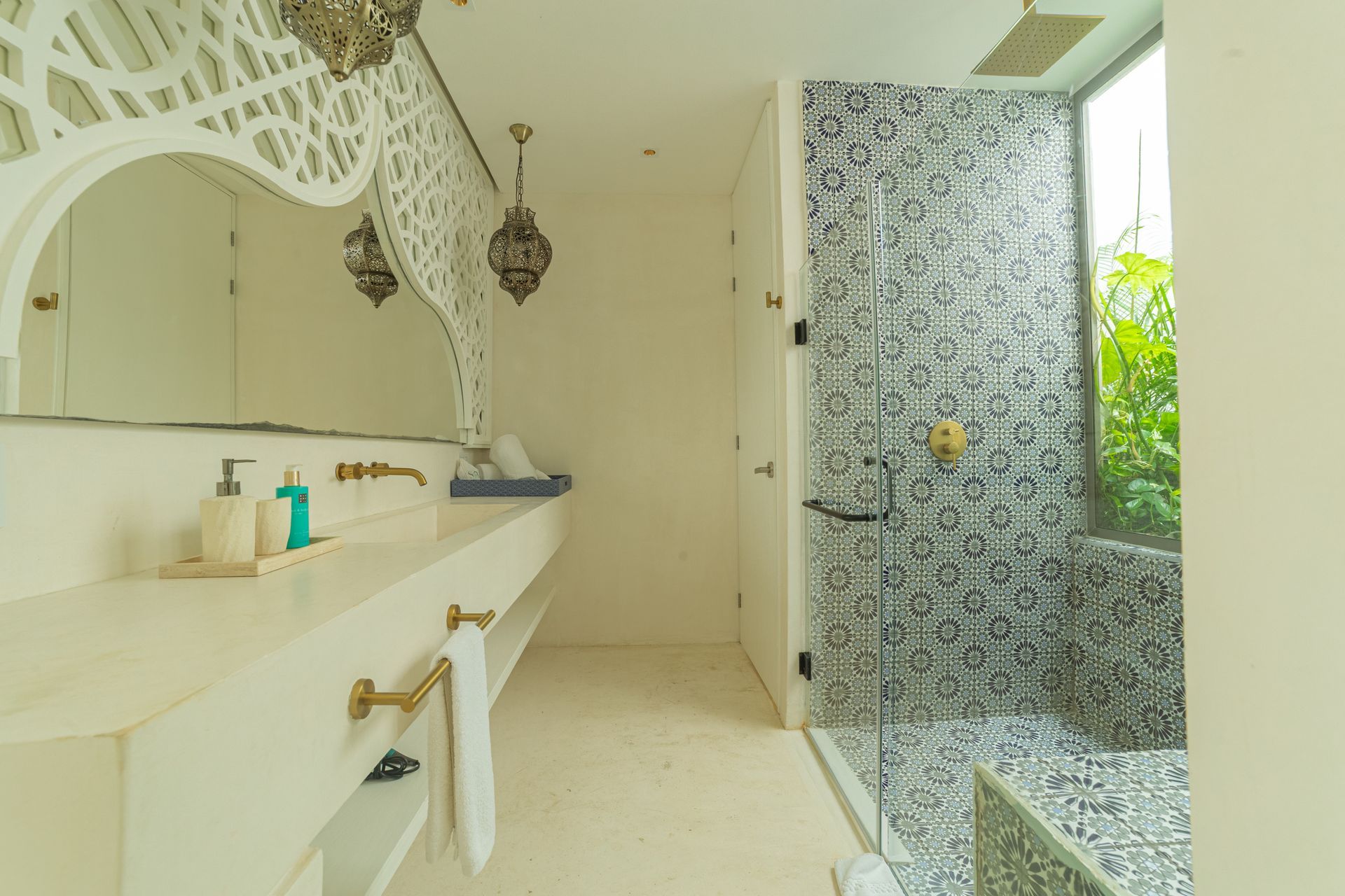Baño blanco con ducha estampada, encimera larga, espejo decorativo y accesorios dorados.