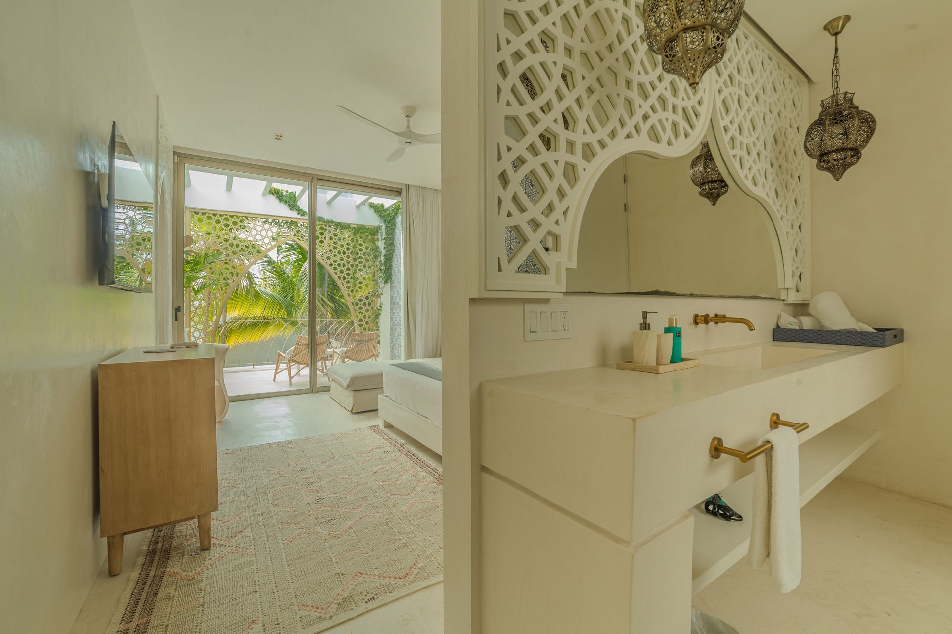 Baño con espejo ornamentado, lavabo blanco y puerta de entrada a una habitación luminosa con árboles visibles.