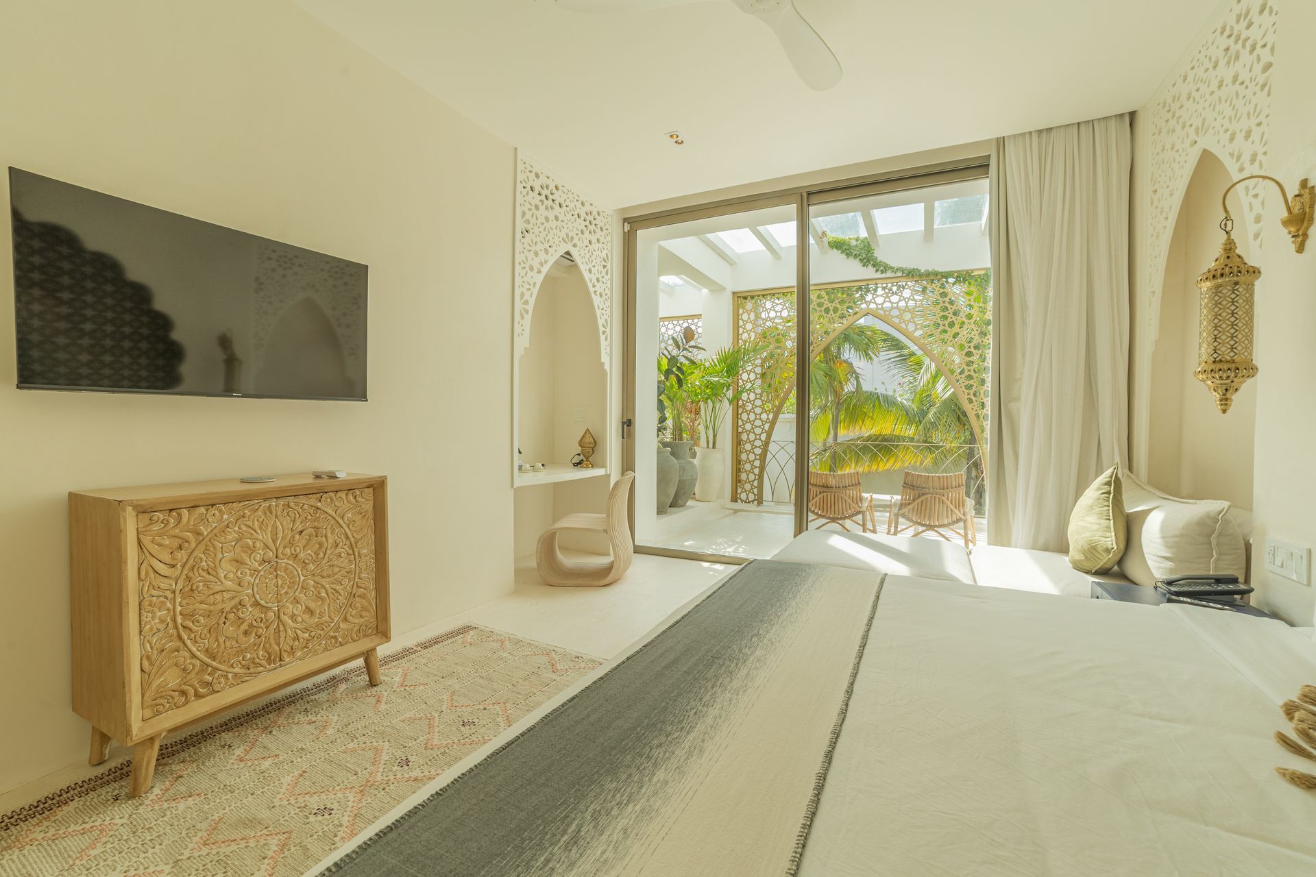 Dormitorio luminoso con muebles de madera tallada, gran ventanal a patio con plantas y tonos neutros.
