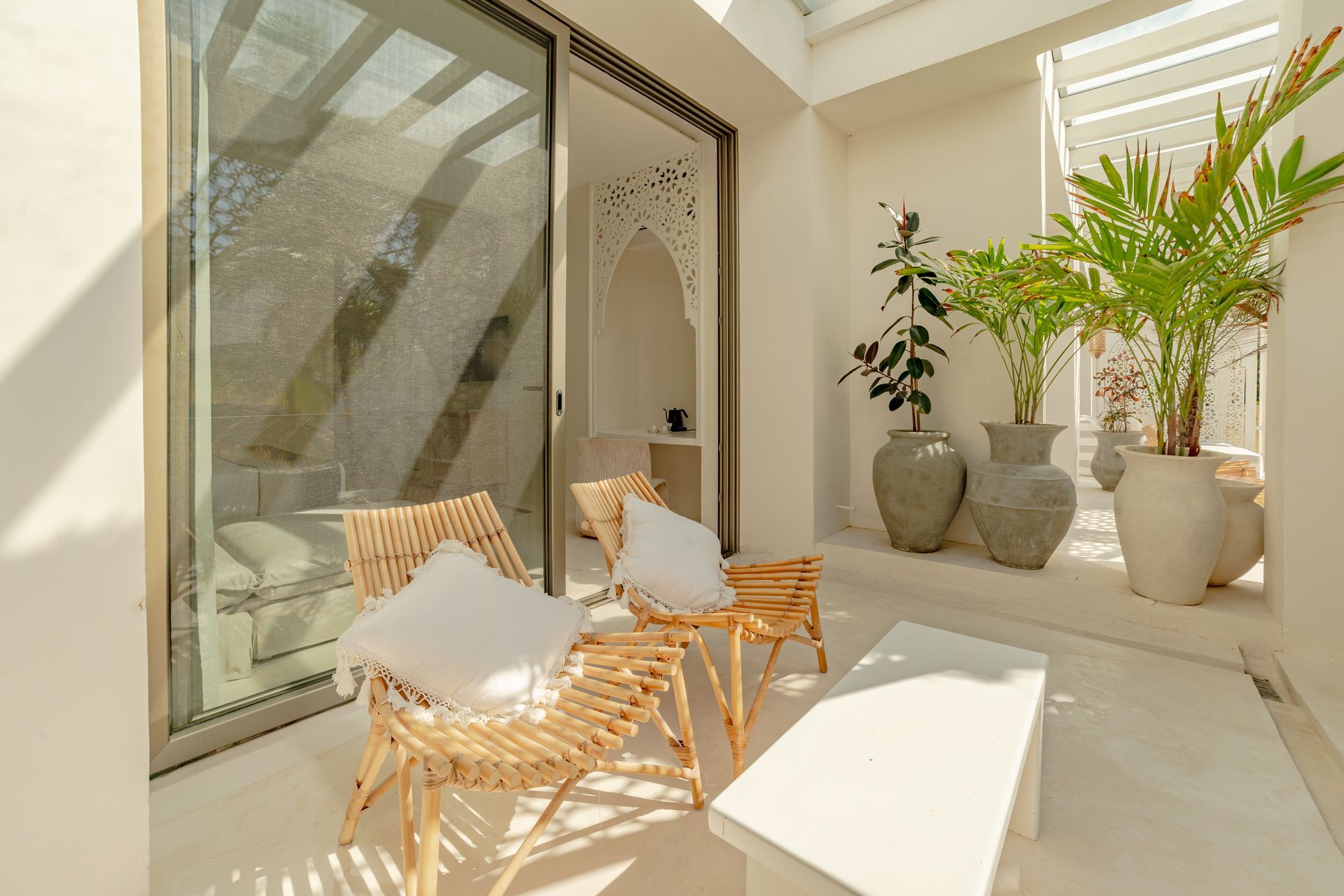 Patio blanco brillante con sillas tejidas, plantas en macetas y puerta corrediza de vidrio.